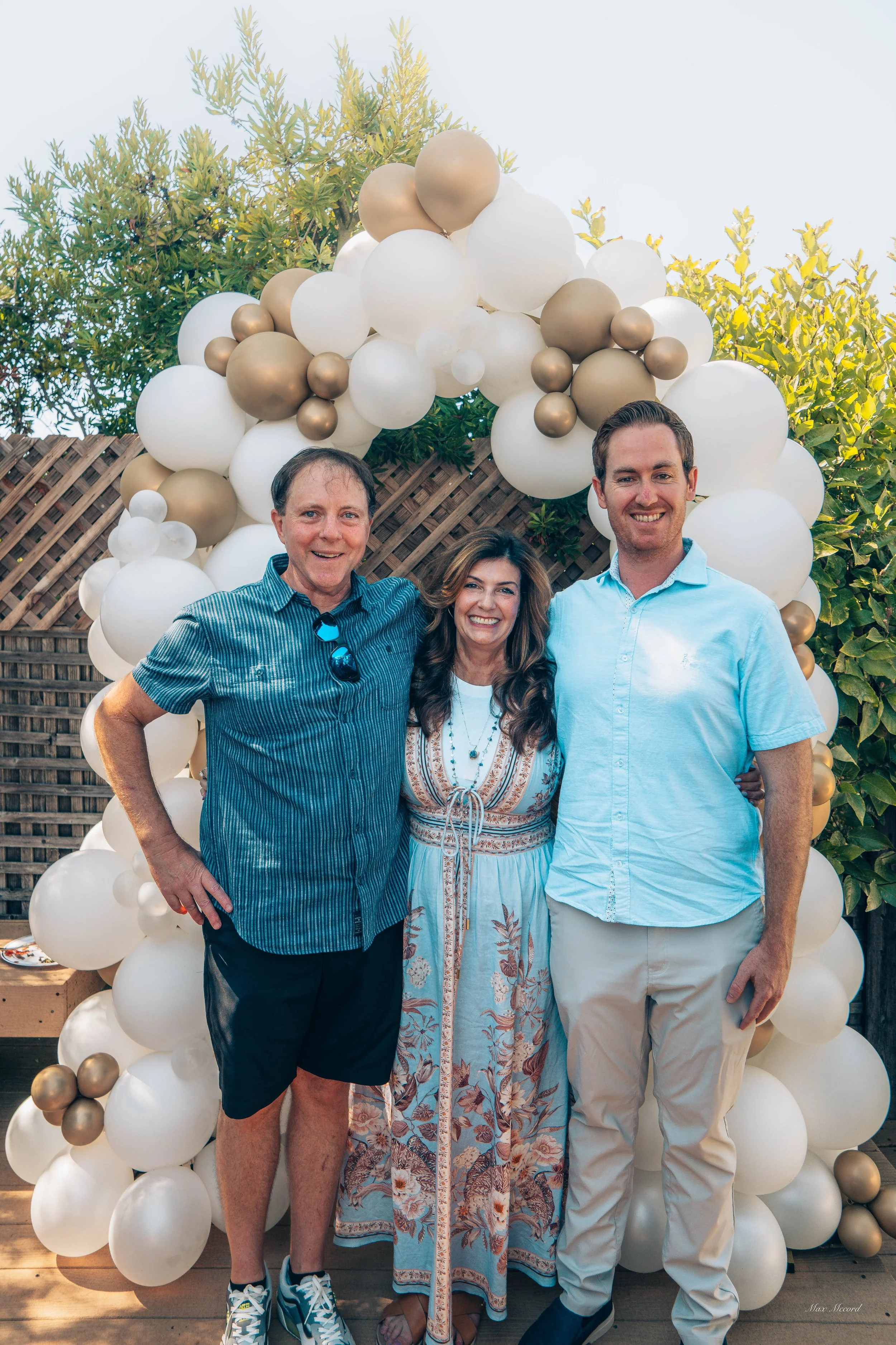Three people smiling in front of a balloon arch at an outdoor celebration.