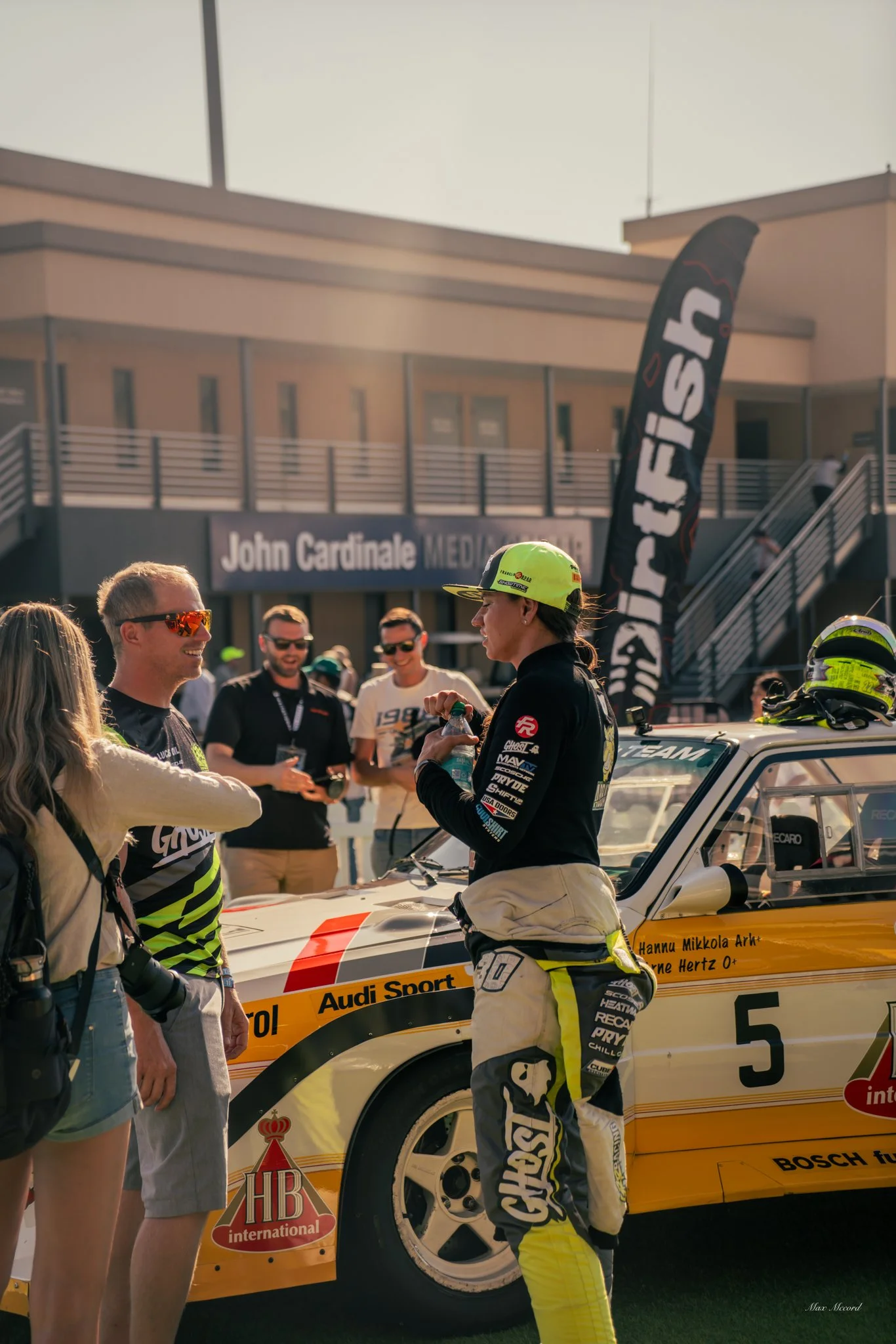 A woman race car driver in racing gear talking to a man and woman with a yellow race car in the background, at what appears to be a racing event.