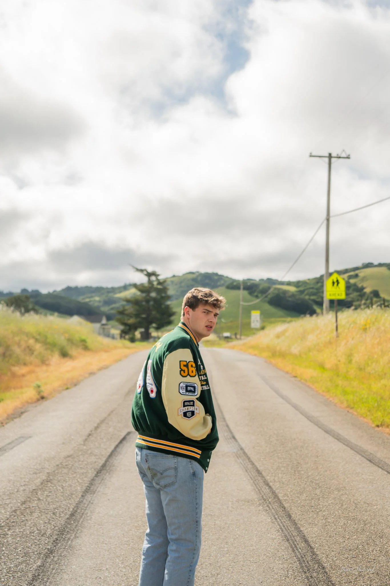 A young man with curly hair wearing a green and beige varsity jacket and light blue jeans stands on the middle of a rural road, looking over his shoulder. The road is flanked by grassy fields and hills, with power poles and a pedestrian crossing sign