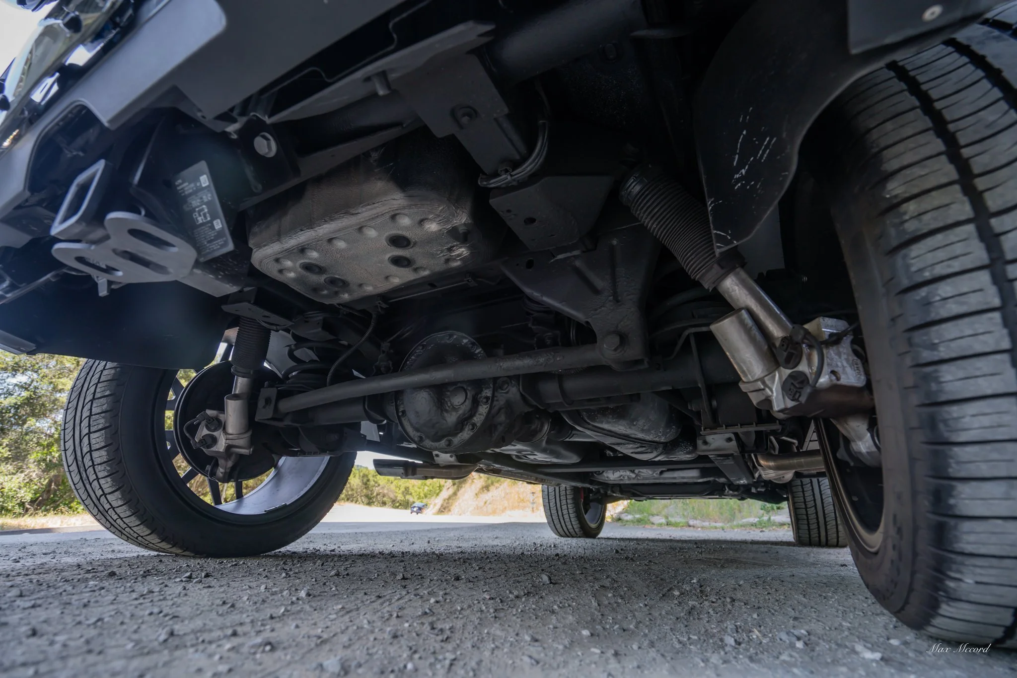 Underneath view of a car shows suspension, driveshaft, and tires on a gravel road.