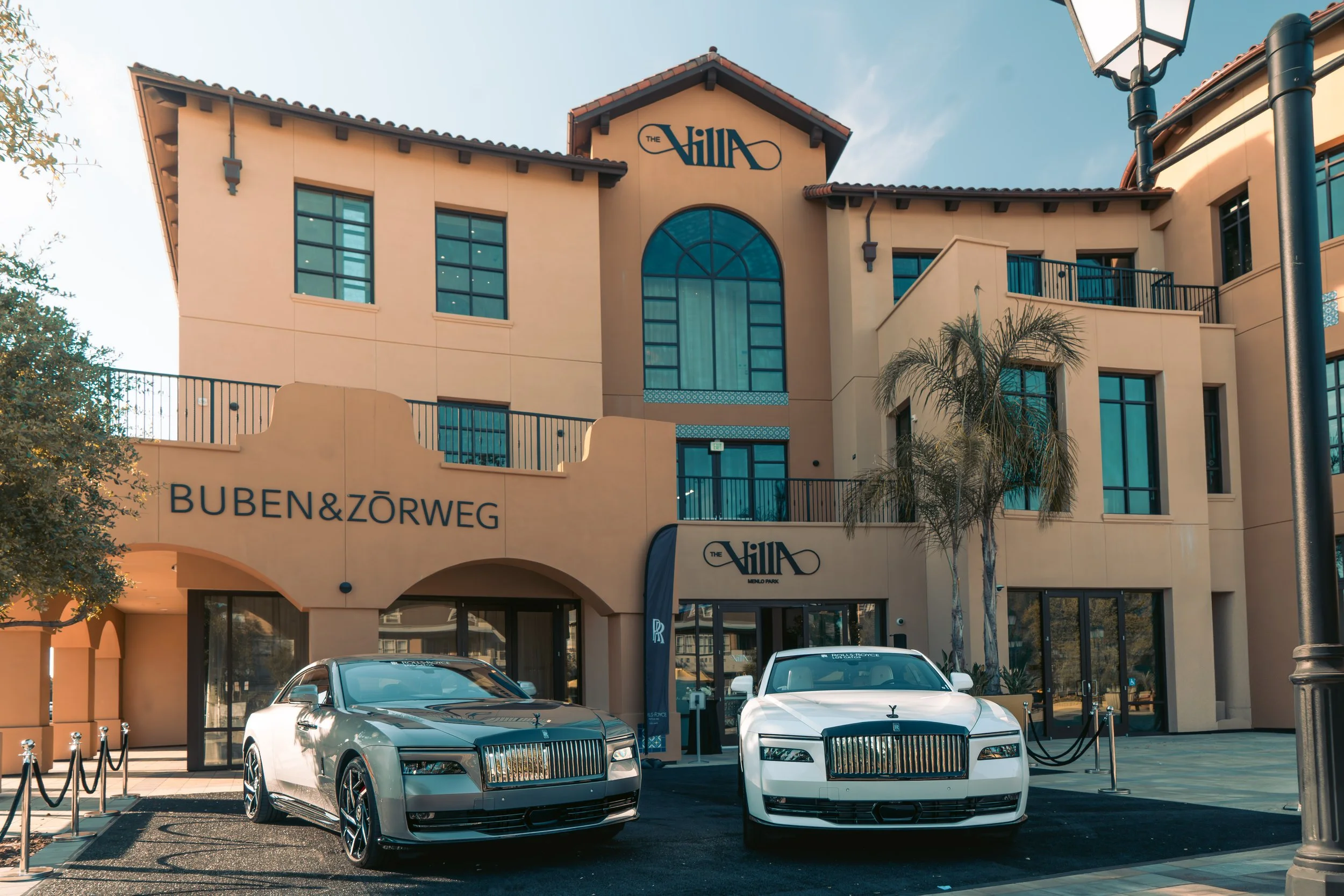 Luxury building with two Rolls-Royce cars parked in front, one silver and one white, outside a high-end shopping center or hotel named The Villa. The building features large windows and palm trees.