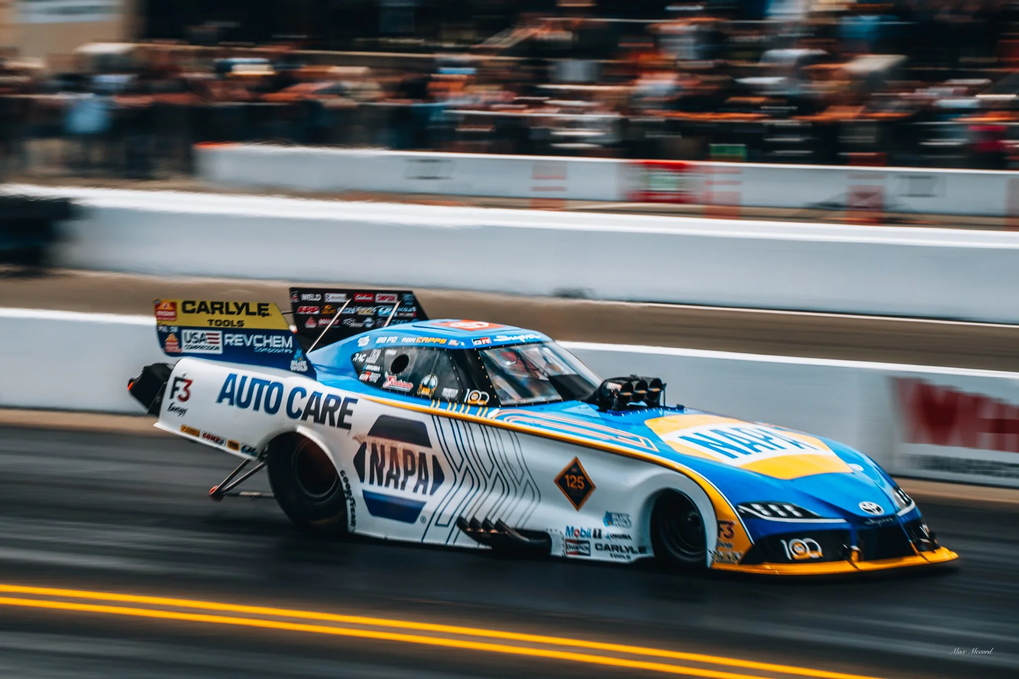 A blue and white NASCAR racing car with various sponsor logos, including NAPA AutoCare, Carlyle Tools, and RevChem, speeding on a race track with spectators blurred in the background.