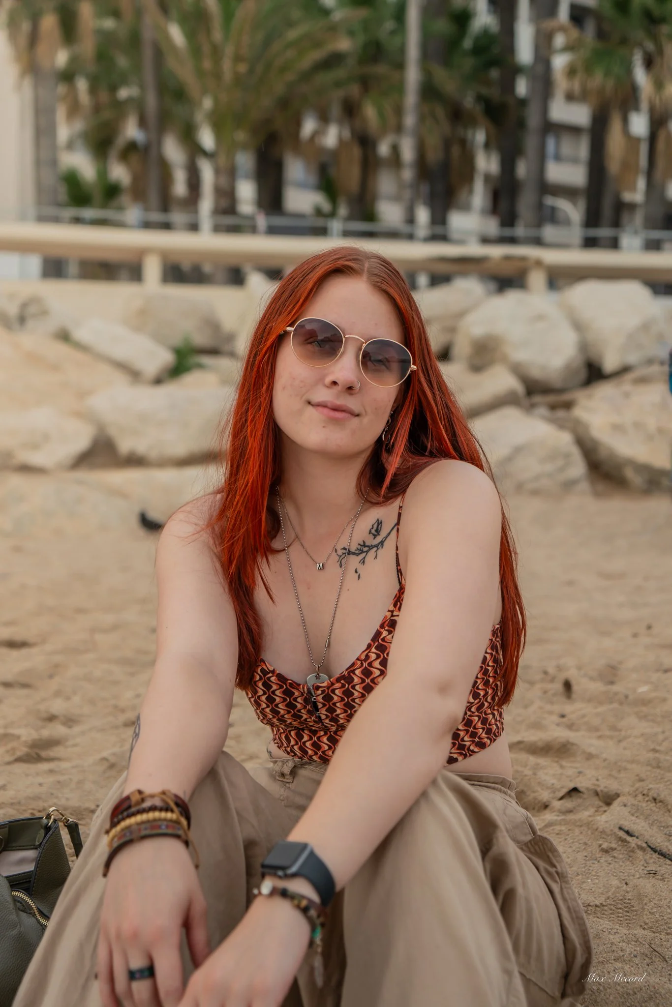 A woman with red hair and sunglasses sitting on the beach with a rock formation behind her.