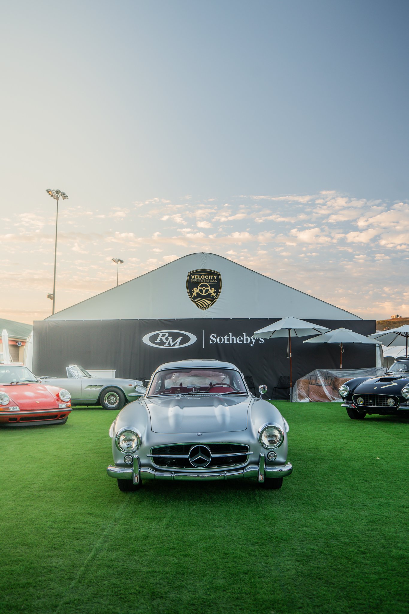 Front view of a silver vintage Mercedes-Benz car parked on green grass at a car show, with other classic cars and a large tent in the background.