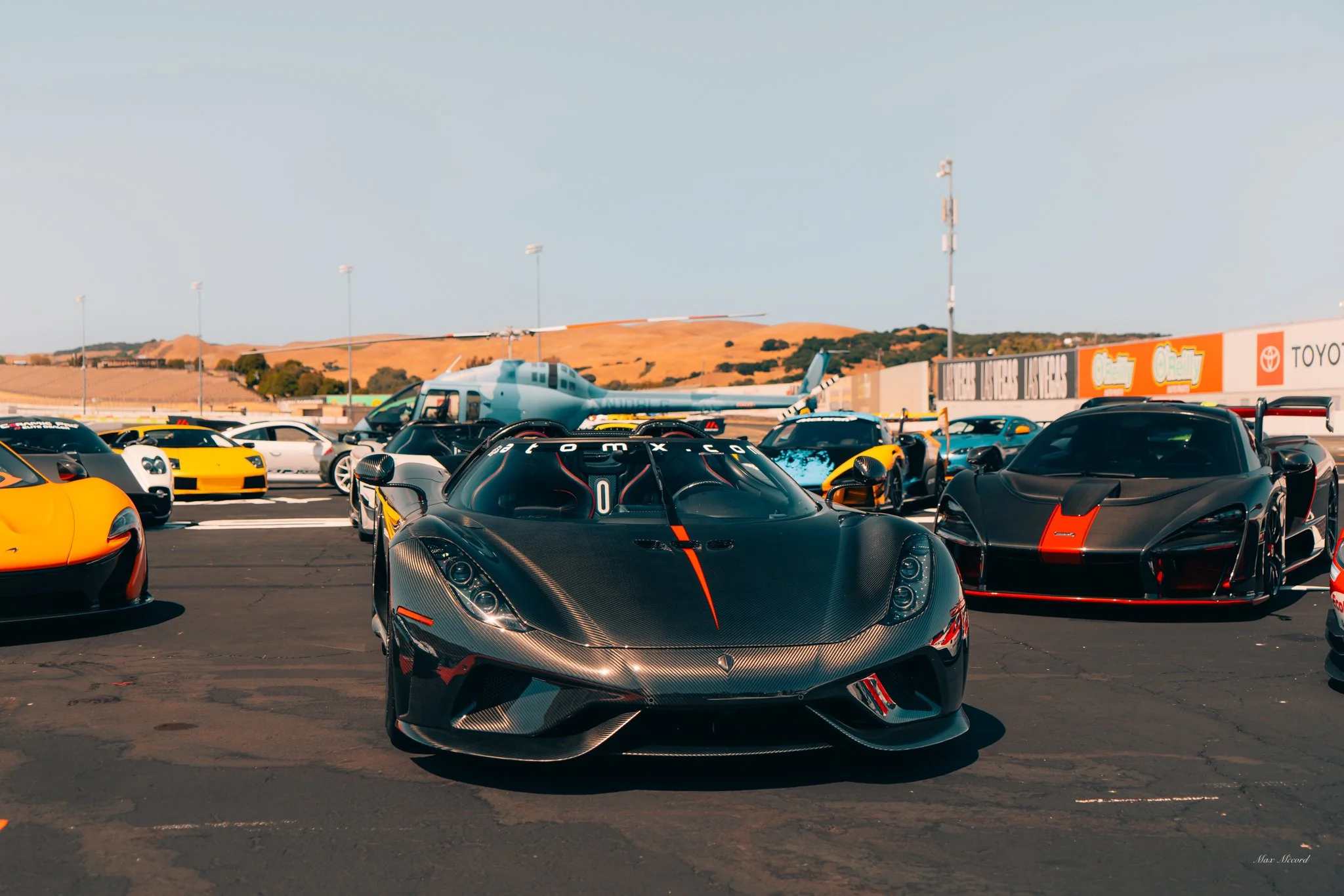 Multiple high-performance sports cars, including a black carbon fiber McLaren with red accents and a black sports car with a red stripe, parked in a racing pit lane with a helicopter in the background and hills in the distance.