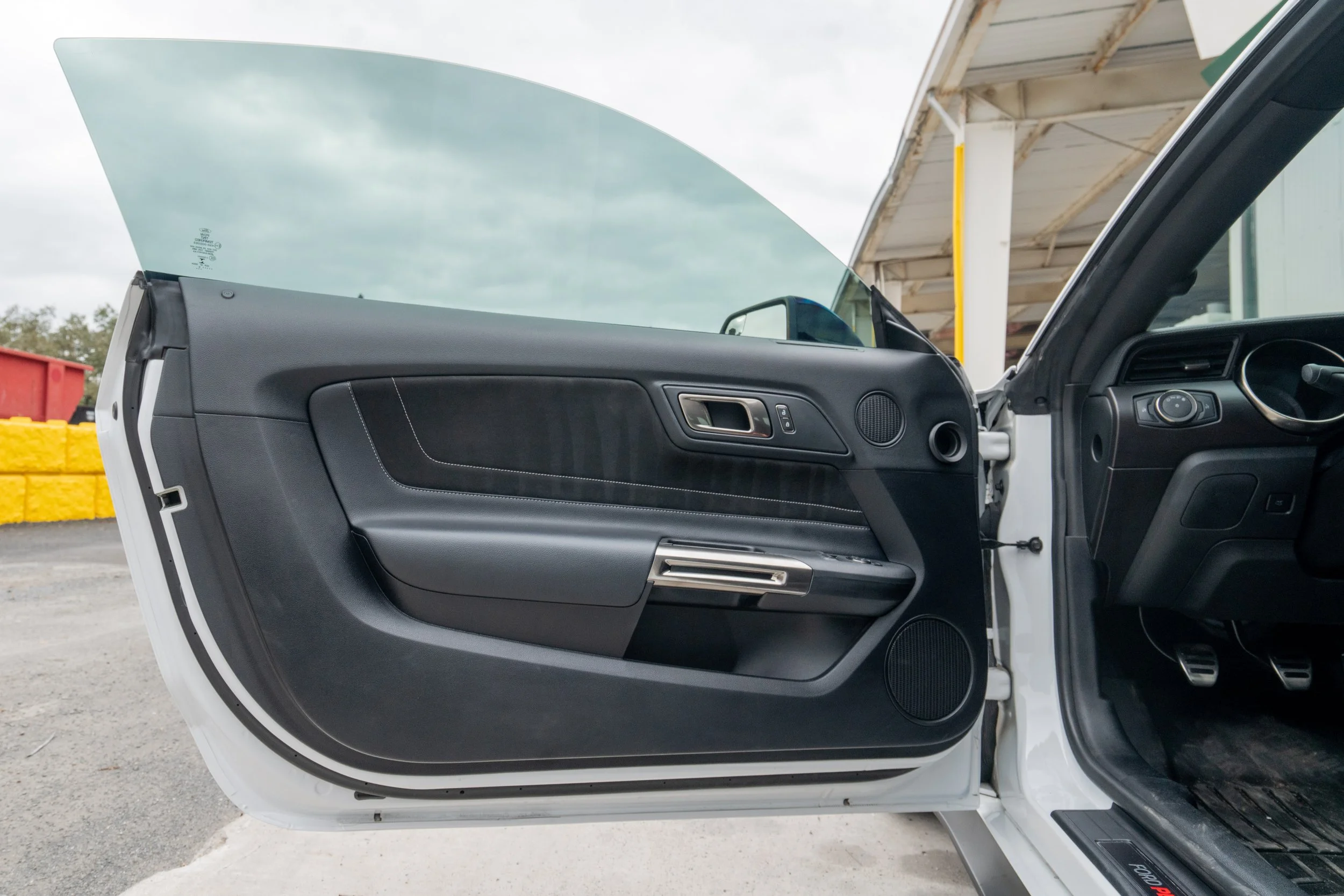 Open car door showing black dashboard and interior, with view of pedals and seat, parked outside near a building with a covered structure and colorful barriers.