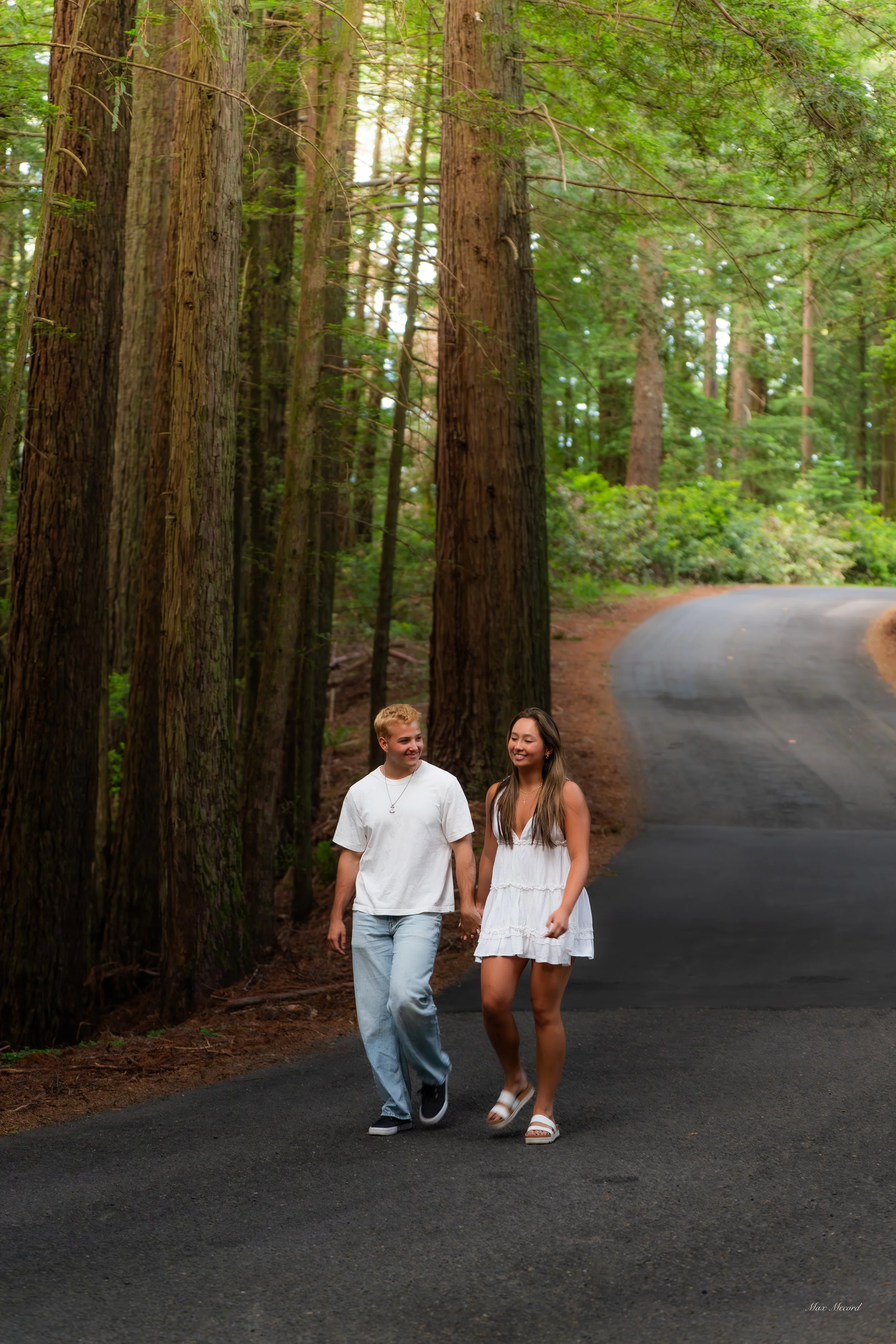A young man and woman walk hand-in-hand along a winding forest trail, surrounded by tall trees with green leaves.