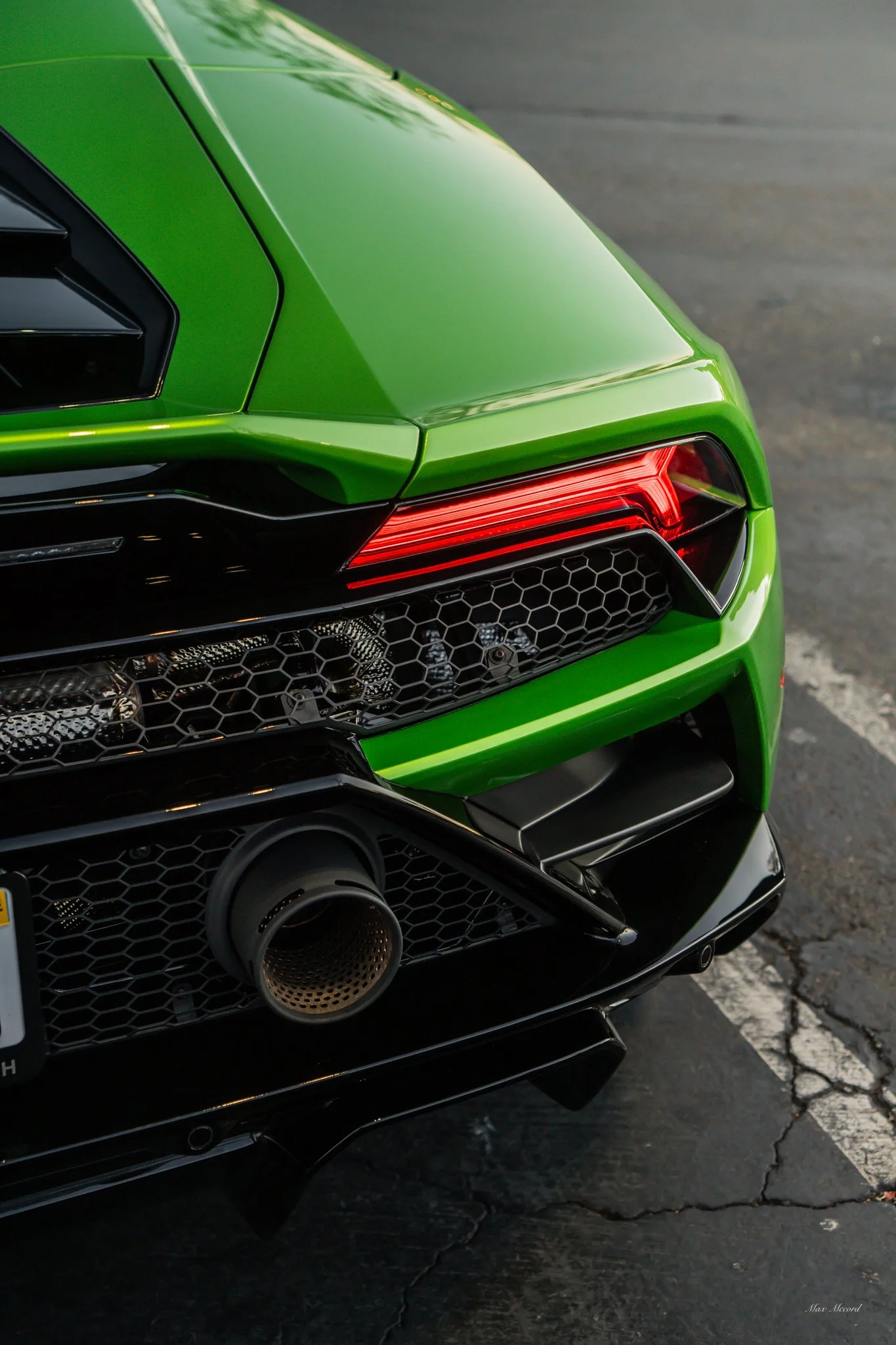 Close-up of the rear end of a bright green sports car with a black honeycomb grille, red tail lights, and a prominent exhaust pipe with a metallic finish, parked on a cracked asphalt surface.