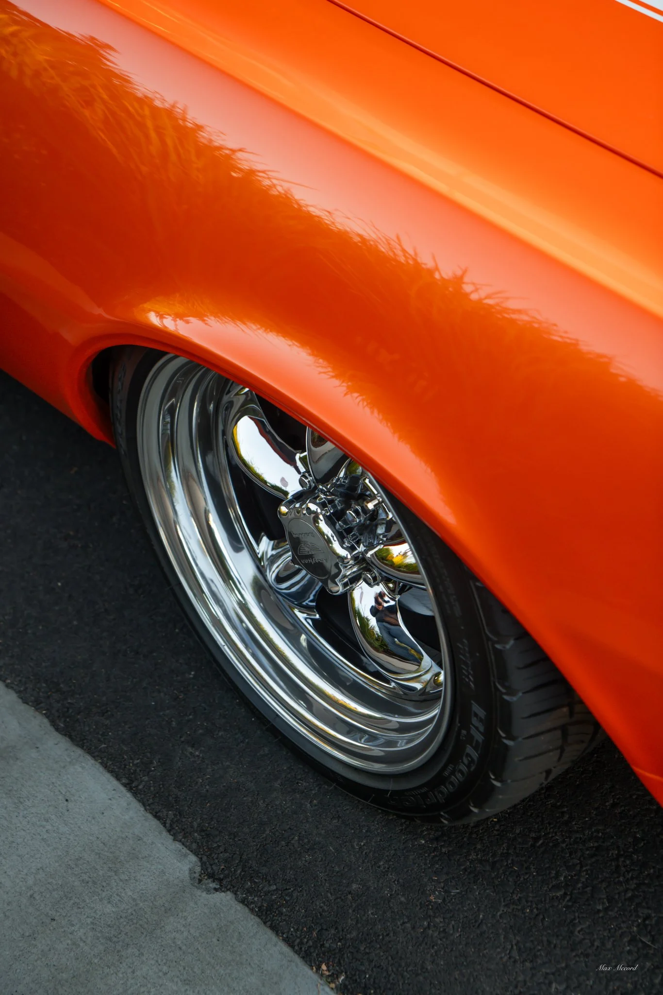 Close-up of an orange car with shiny chrome wheel and tire on a black asphalt surface.