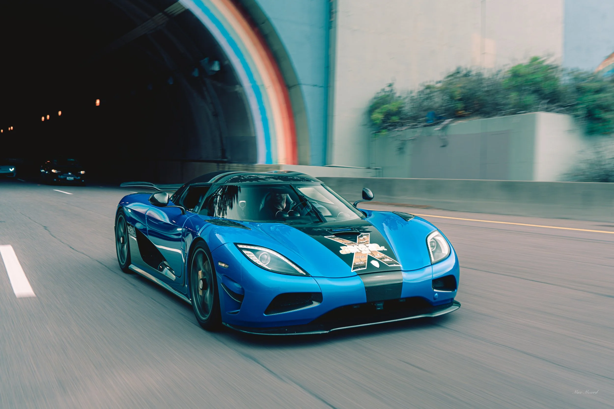 Blue sports car with black racing stripes driving through a tunnel with colorful lights.
