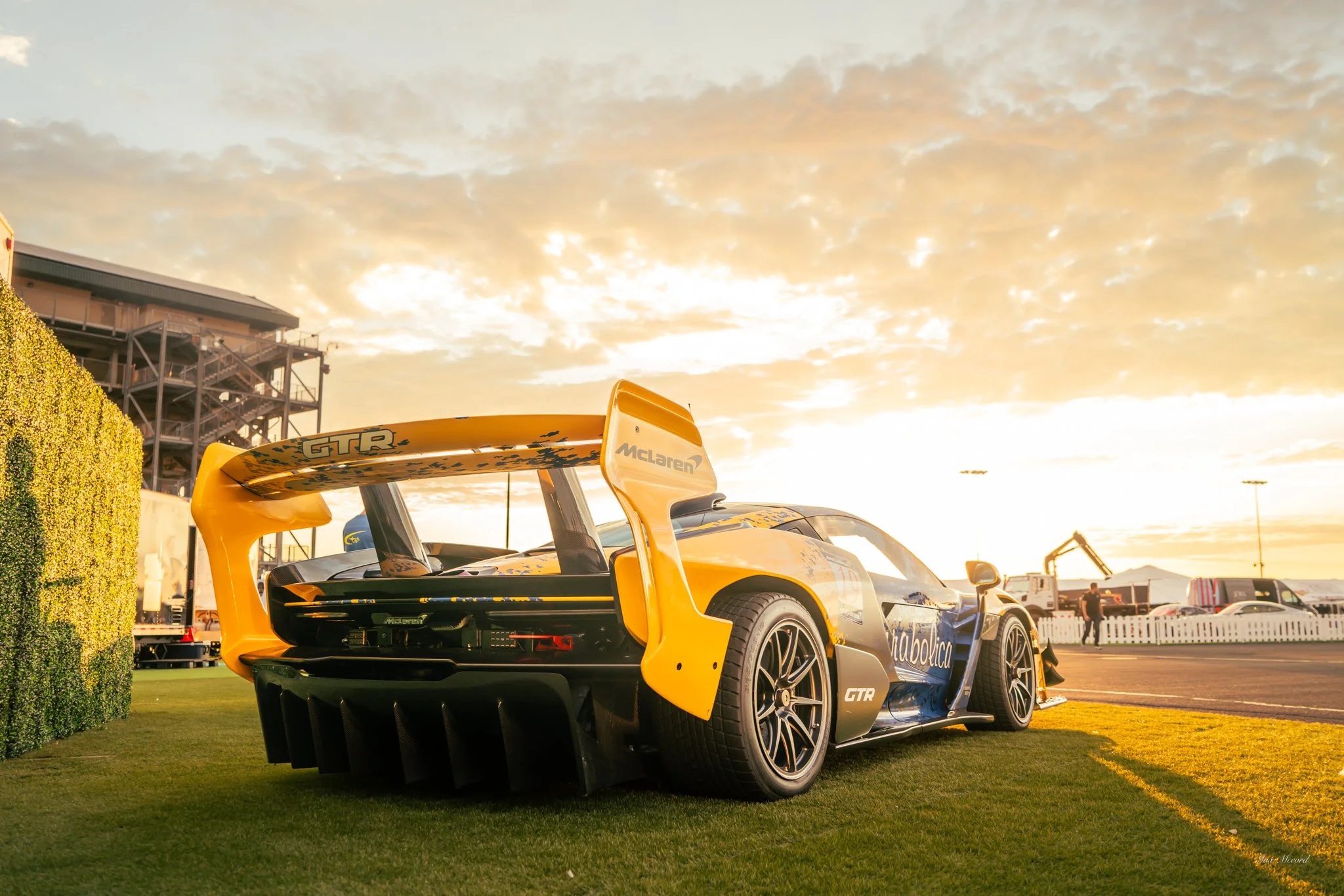 A yellow and black McLaren race car with a large rear wing parked on a grassy area at sunset. The background shows a racing circuit, some construction equipment, and spectators.
