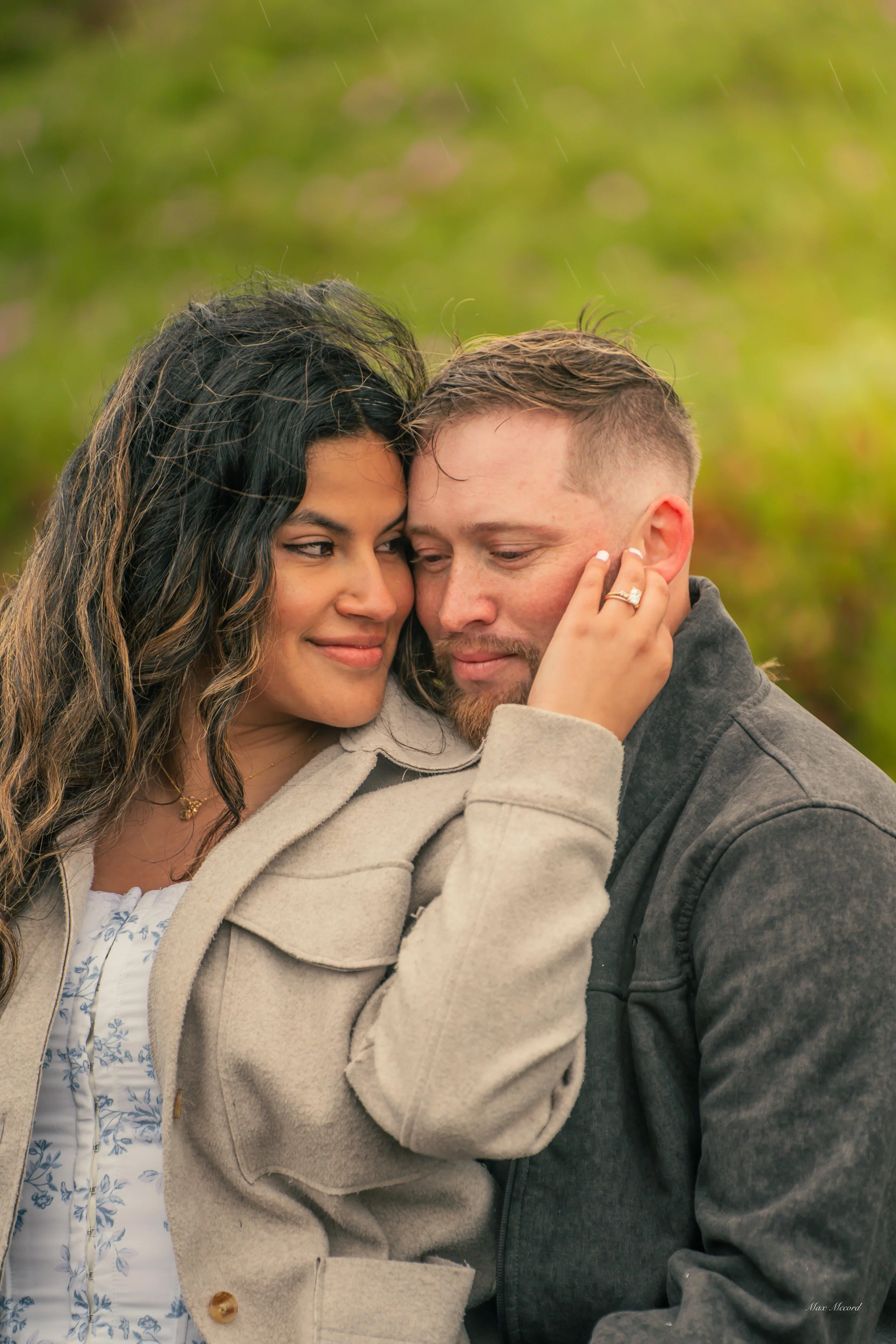 A woman and a man embrace outdoors on a rainy day, with the woman gently touching the man's face and both appearing happy and close.