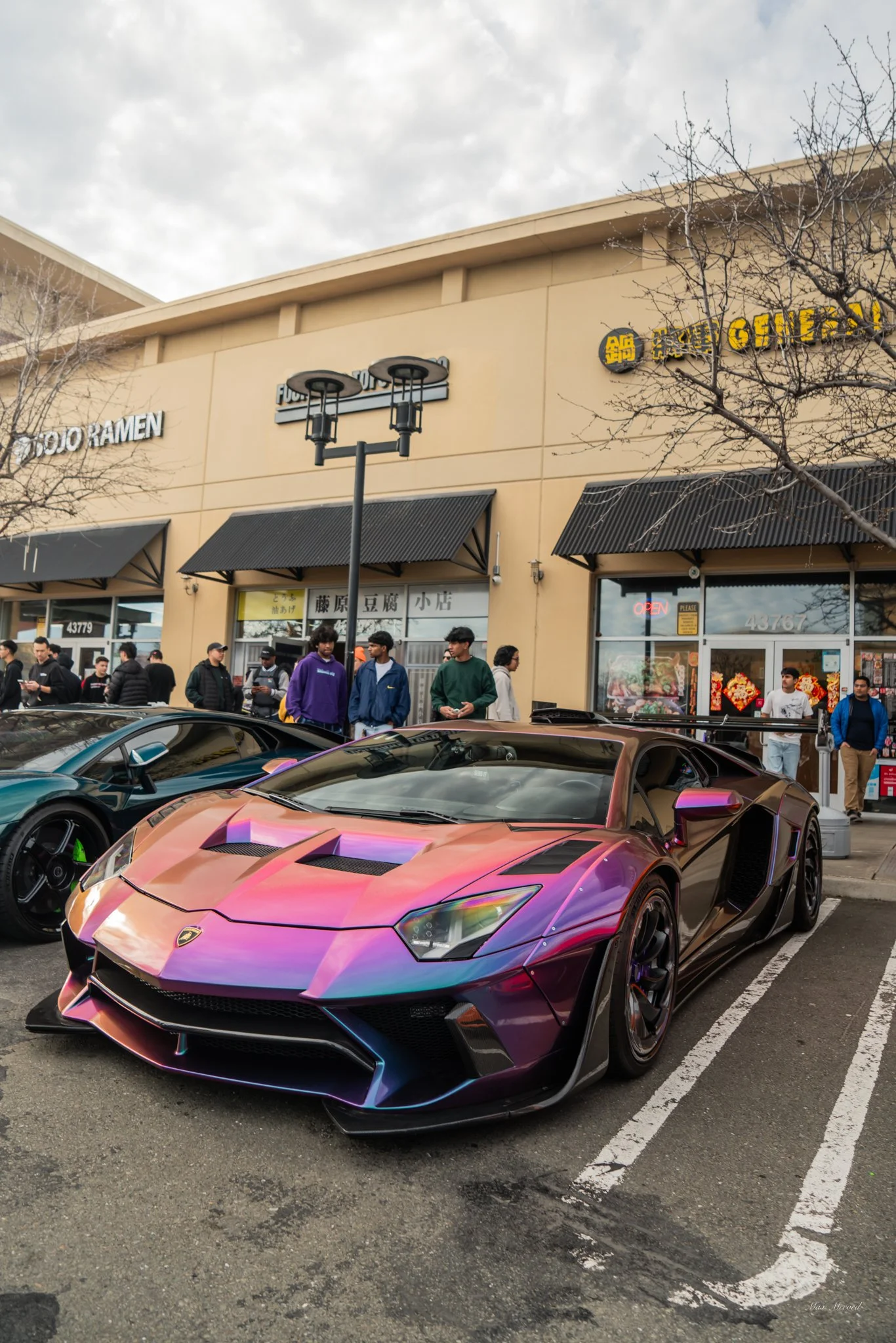 A purple and black Lamborghini sports car parked in front of a shopping center. People are walking and standing in line outside stores. The shopping center has beige walls with black awnings and signs for various businesses.