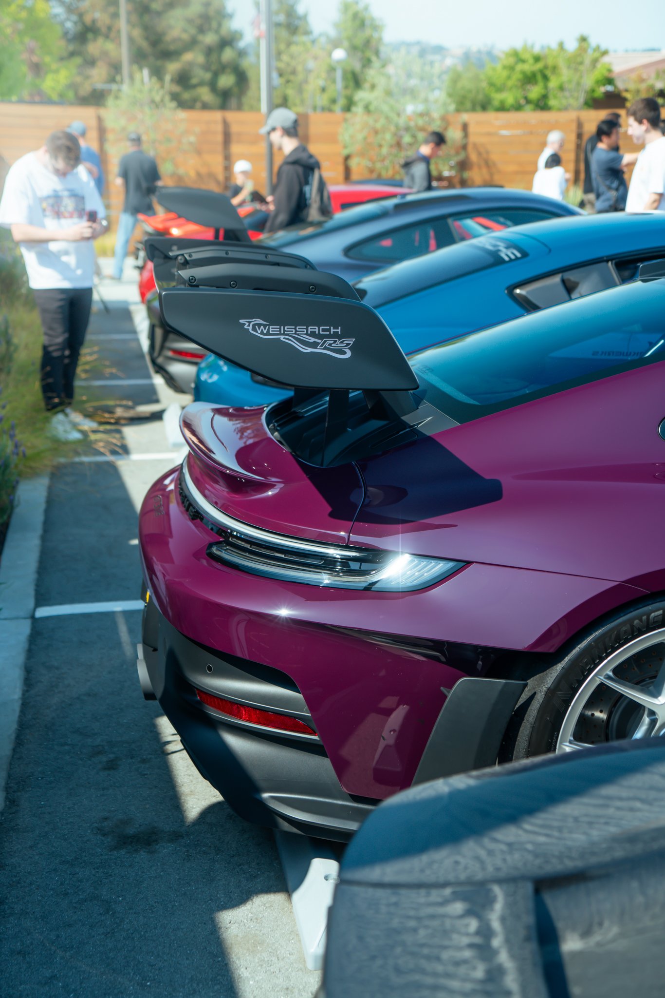 Close-up of the rear of a maroon Porsche 911 with Weissach RS wing, parked among other sports cars at a car show, with people walking around and taking photos in the background.