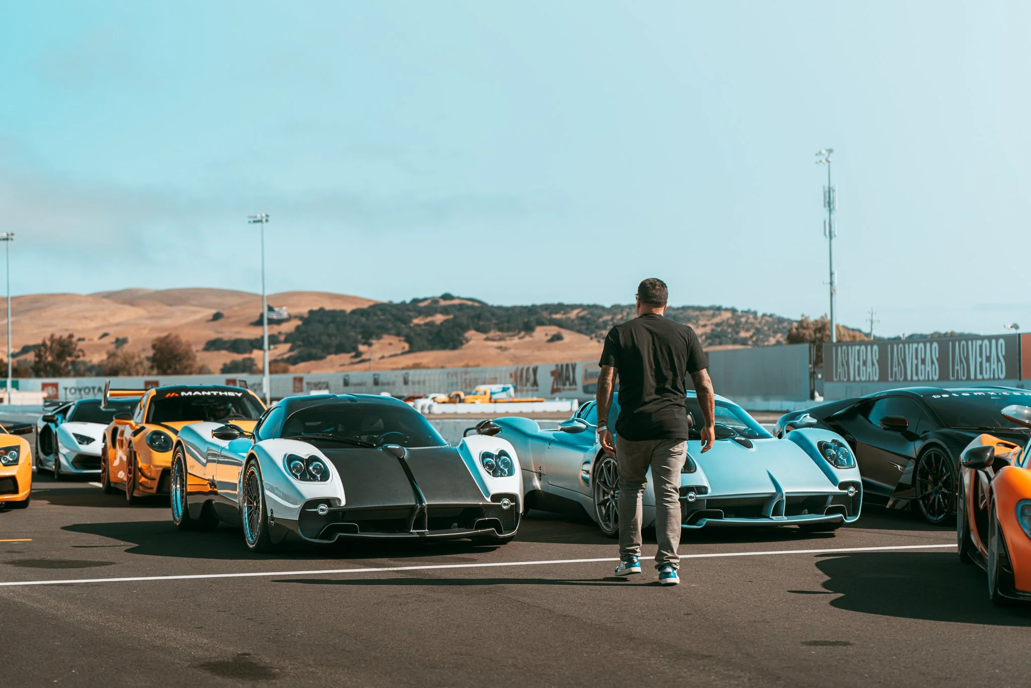 A man walking past luxury sports cars on a racetrack, with hills and a blue sky in the background.