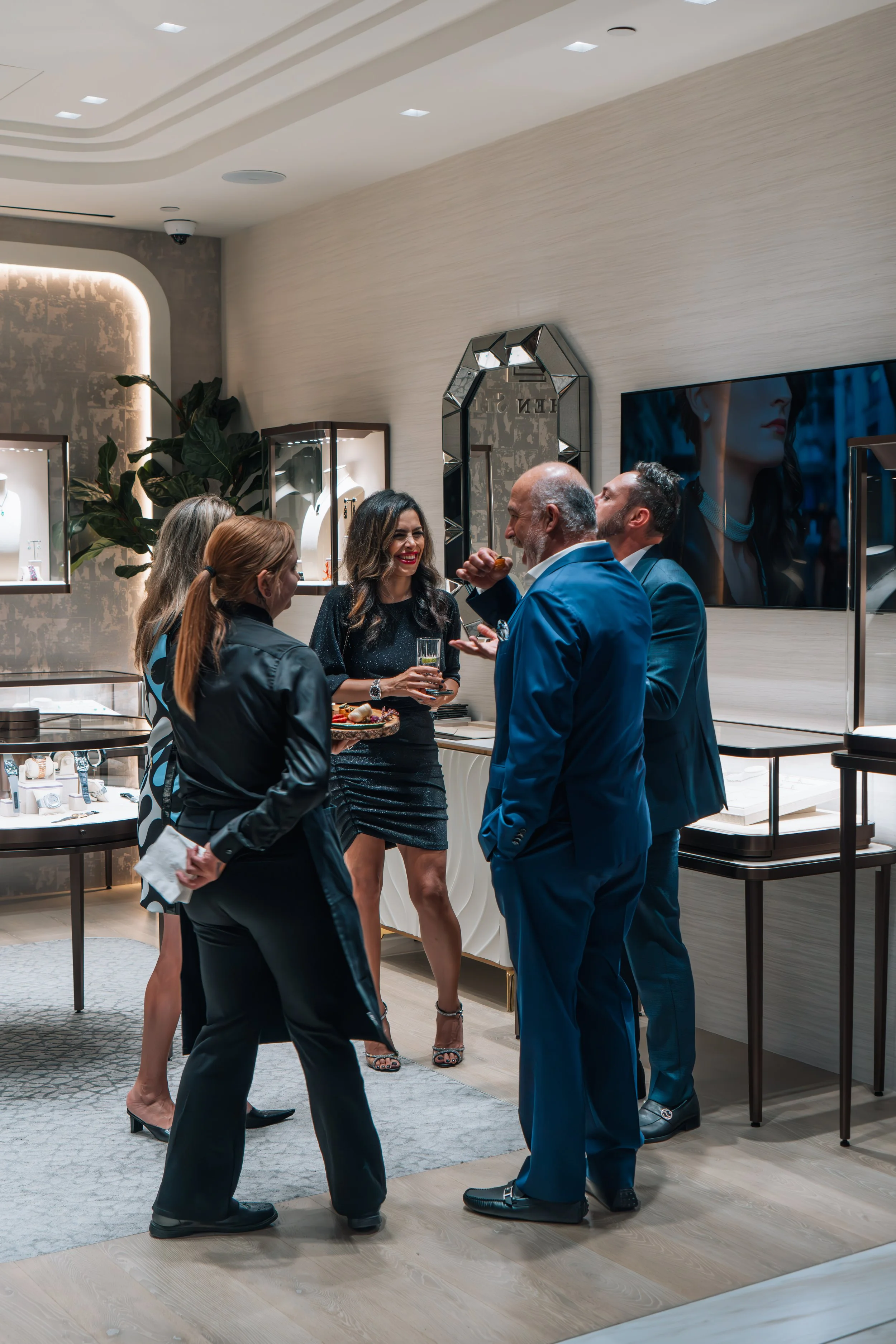 Group of five people in formal attire socializing inside a modern jewelry store, with display cases and a large mirror on the wall.