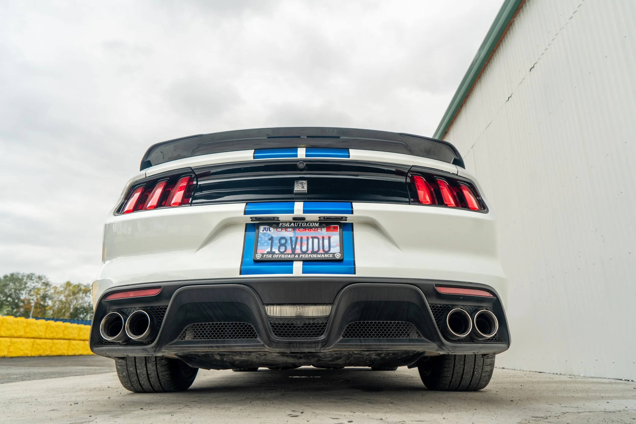 Rear view of a white Ford Mustang performance car with blue racing stripes, dual exhaust pipes, and California license plate.