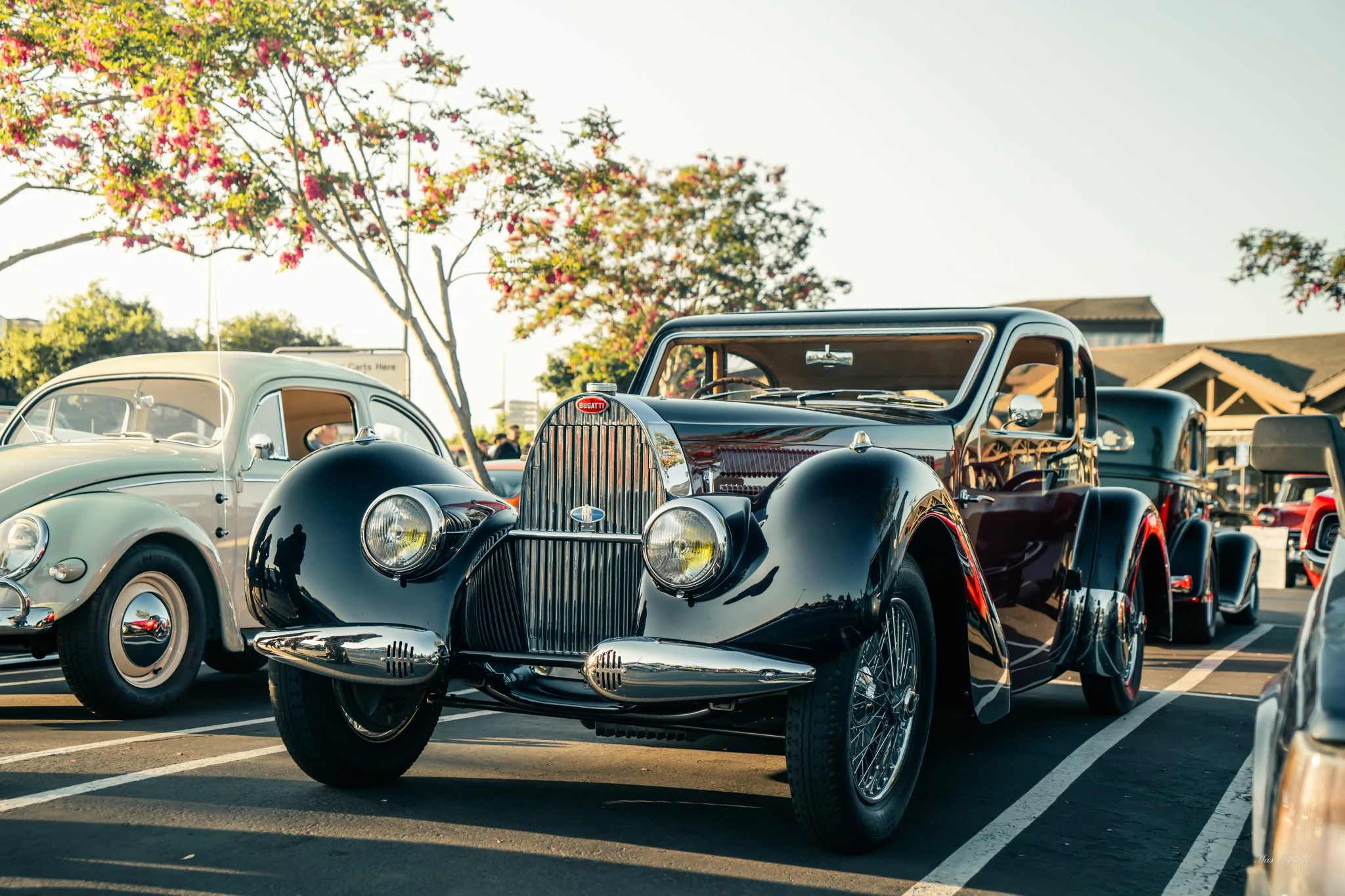 A black vintage Bugatti car parked next to a white classic Volkswagen Beetle at a car show, with trees and other cars in the background during daylight.