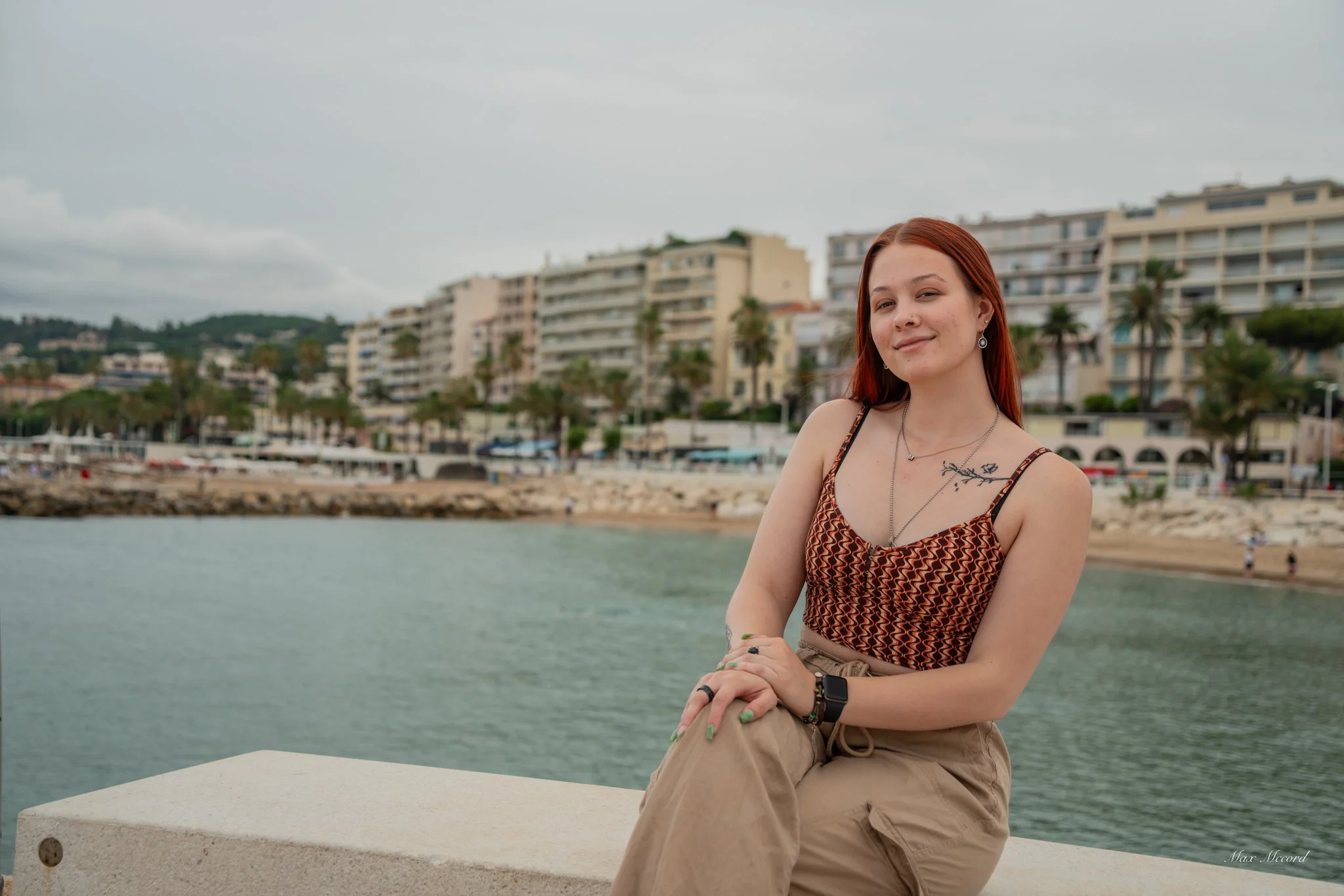 A young woman with red hair sitting on a concrete ledge by the water, with a coastal city and buildings in the background.