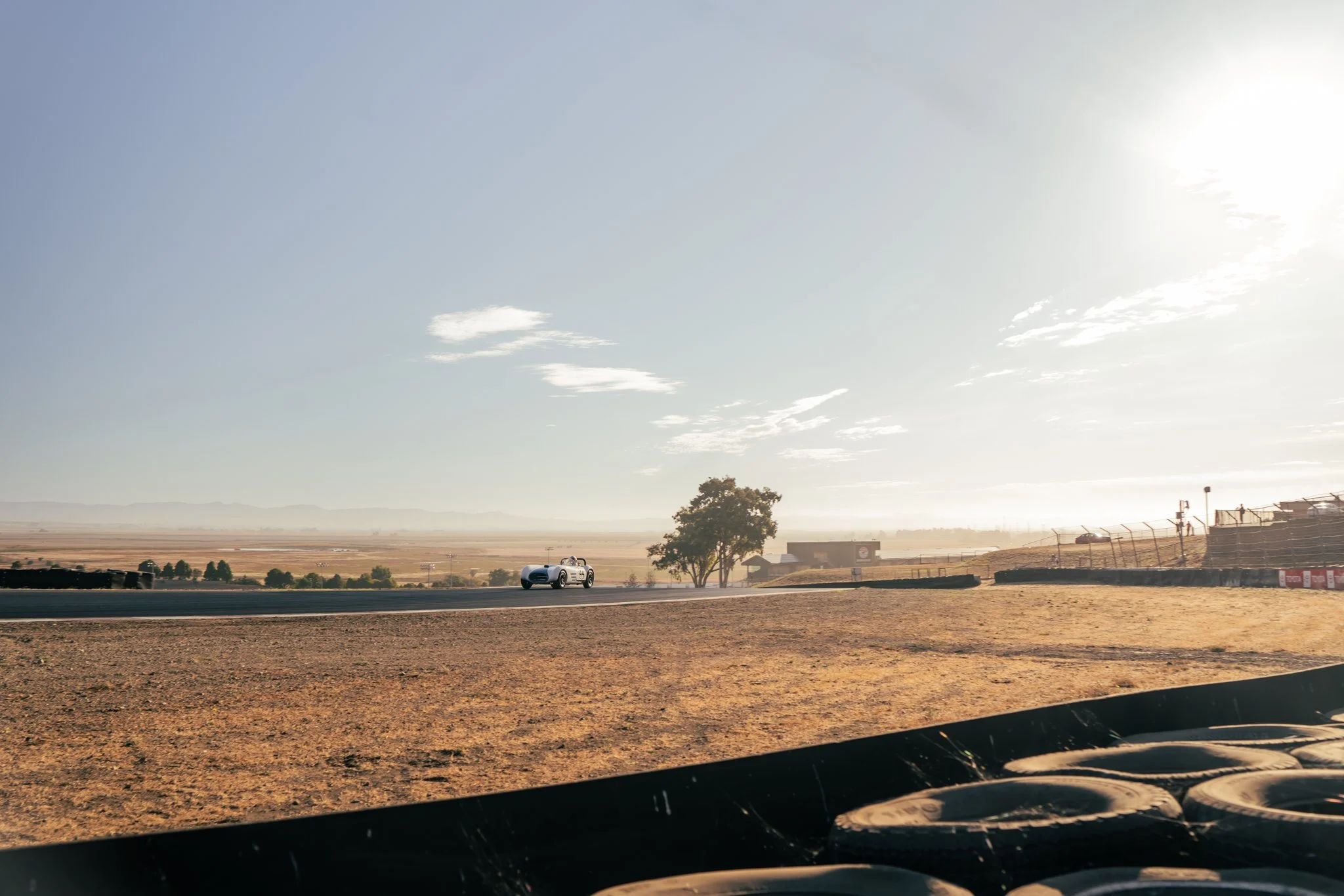 A white race car on a race track in a desert landscape, with sunglasses in the foreground, a tree, and a building in the background under a clear sky.