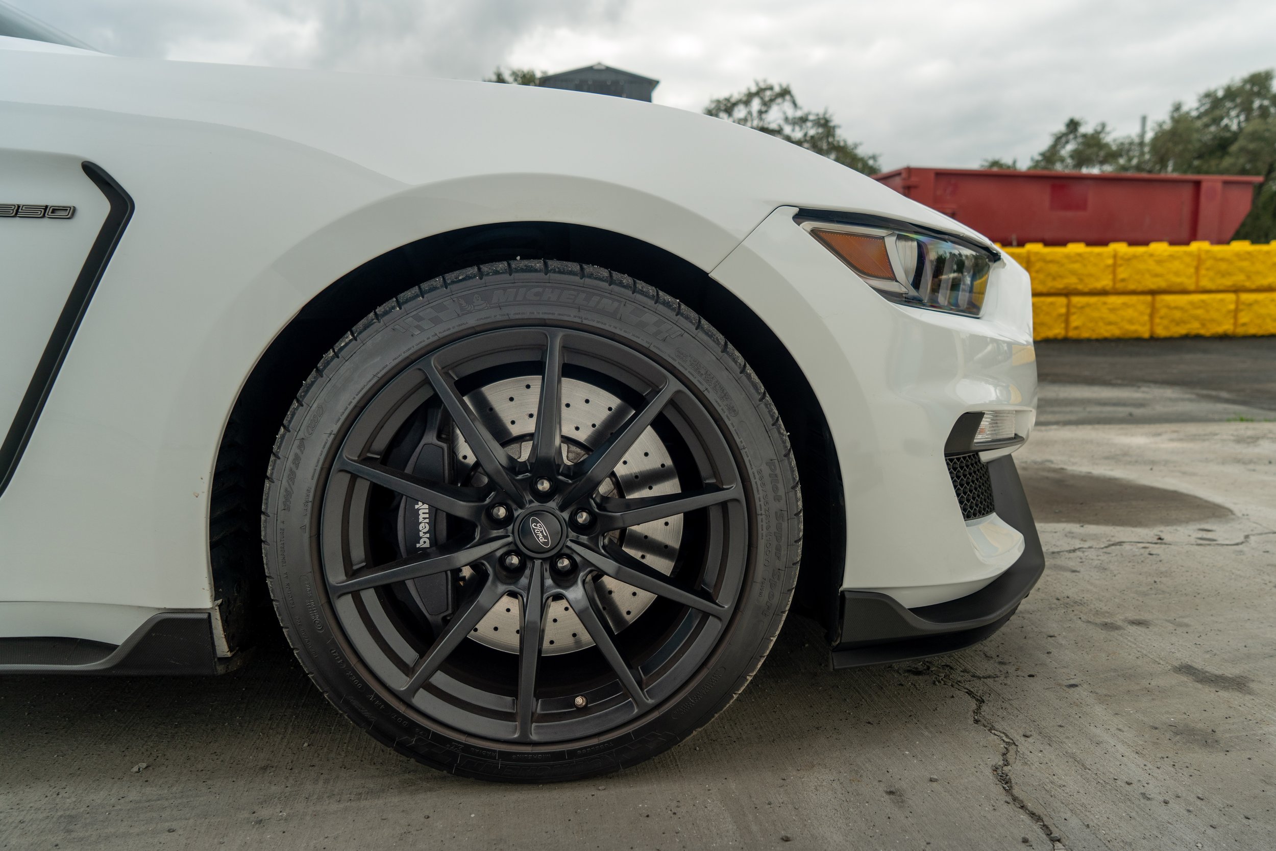 Close-up of the front left side of a white Ford Mustang car, showing a black wheel with a large brake disc and brake caliper, parked on a concrete surface with a yellow/red barrier and cloudy sky in the background.