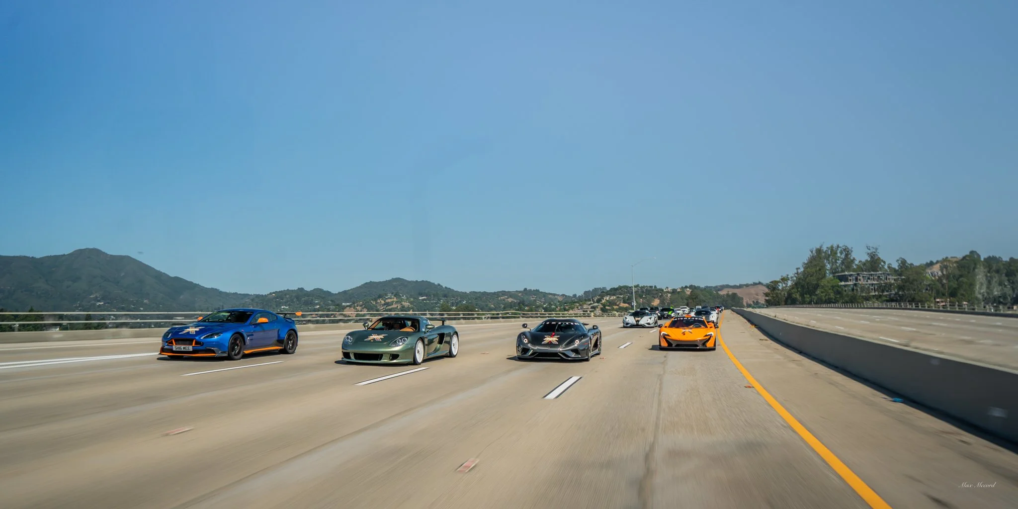 Several sports cars driving on a highway against a backdrop of mountains and blue sky.