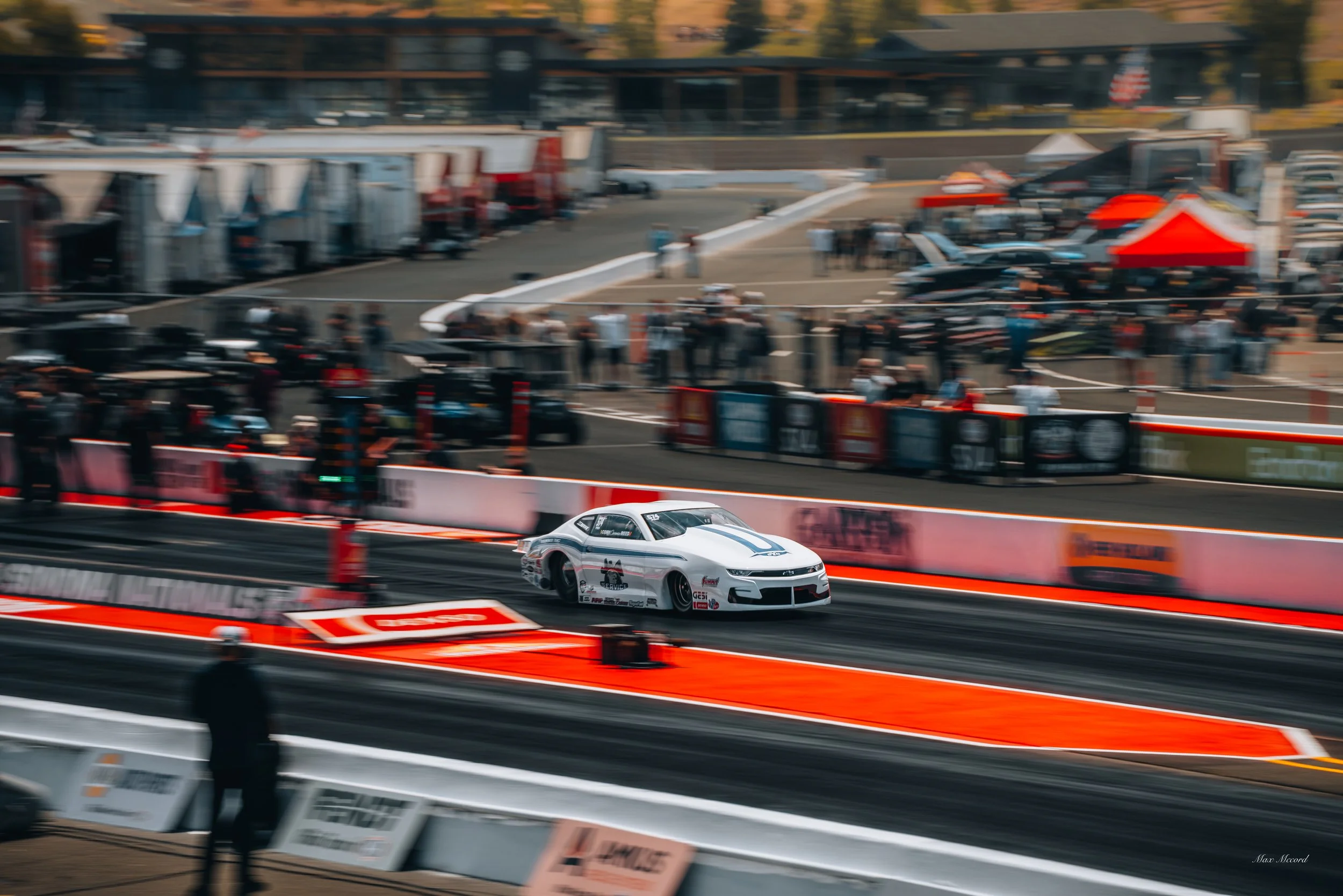 A white race car with blue accents on a drag strip during a racing event, with spectators and tents in the background.