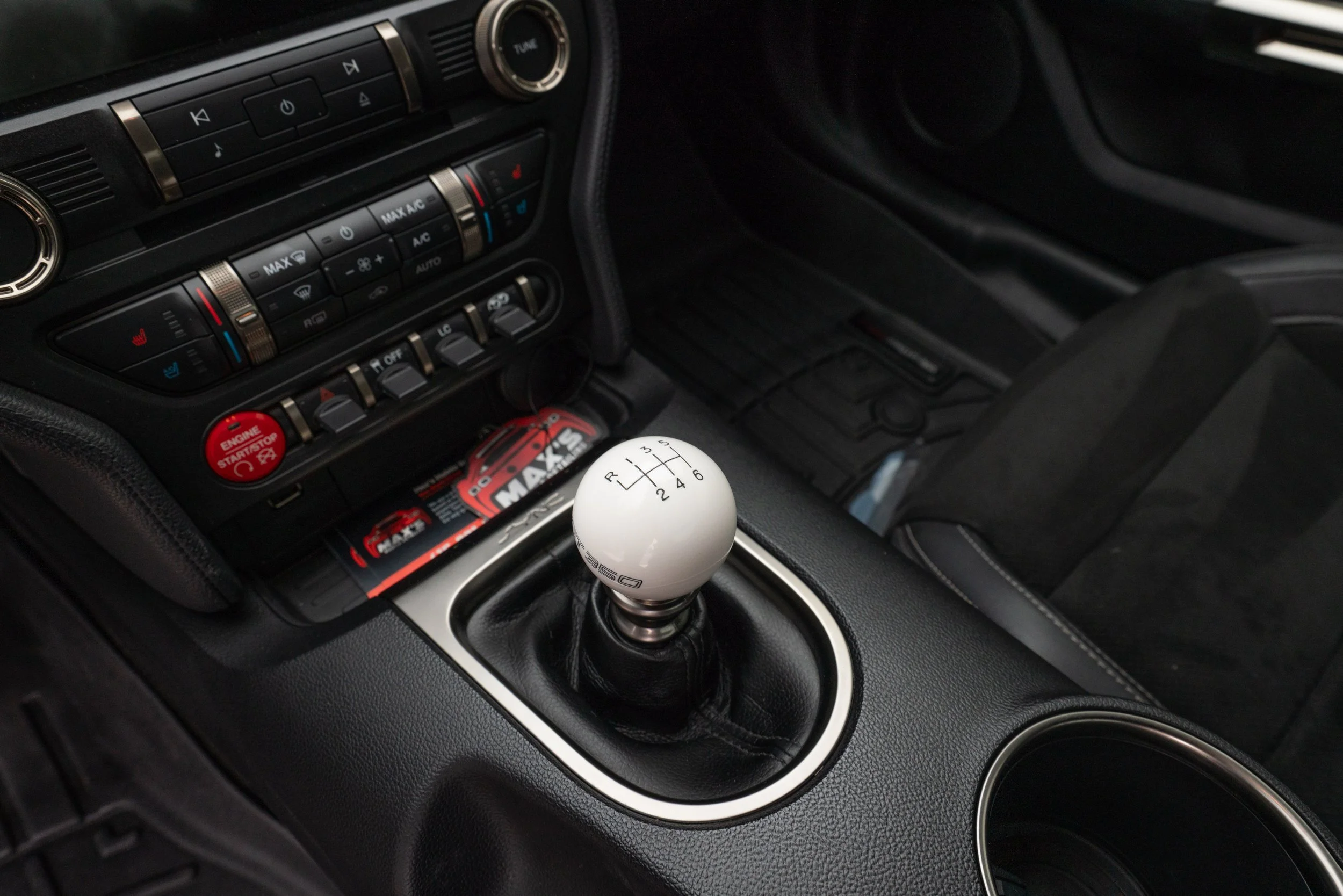 Interior of a car featuring a manual gear shift with a white shift knob, black leather boot, and the center console with buttons and controls.