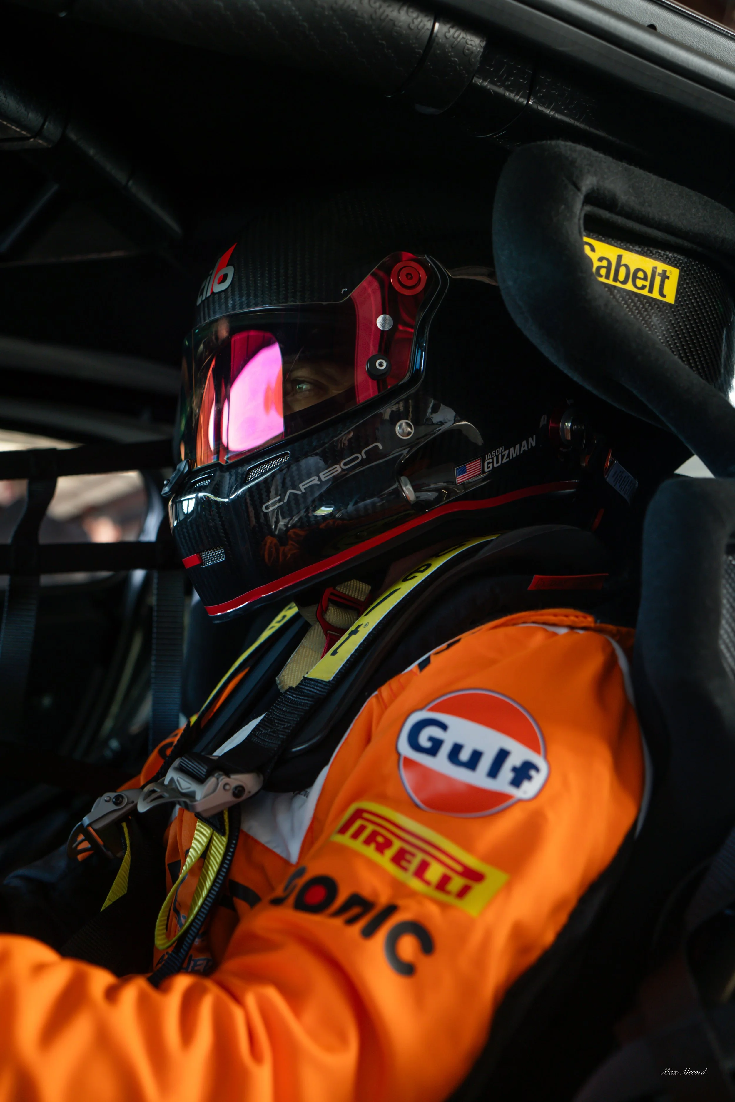 A race car driver in a black helmet with red accents and reflective visor, wearing an orange firesuit with logos including Gulf, Pirelli, and Ion C, seated inside a race car cockpit.