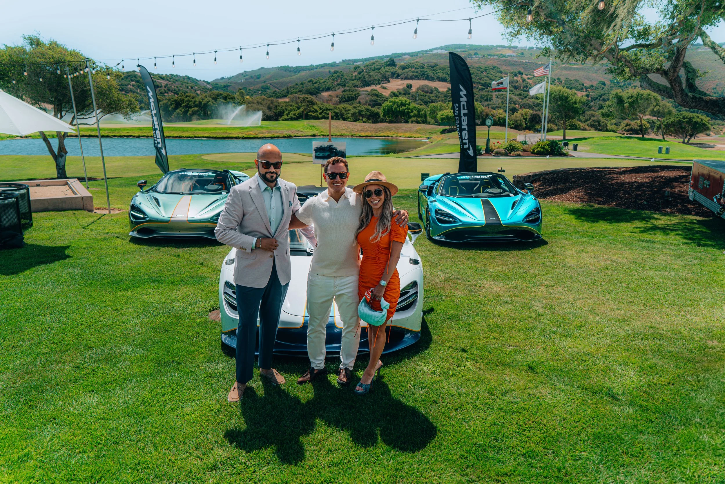 Three people standing in front of luxury sports cars at a car event outdoors, with a pond, hills, and flags in the background.