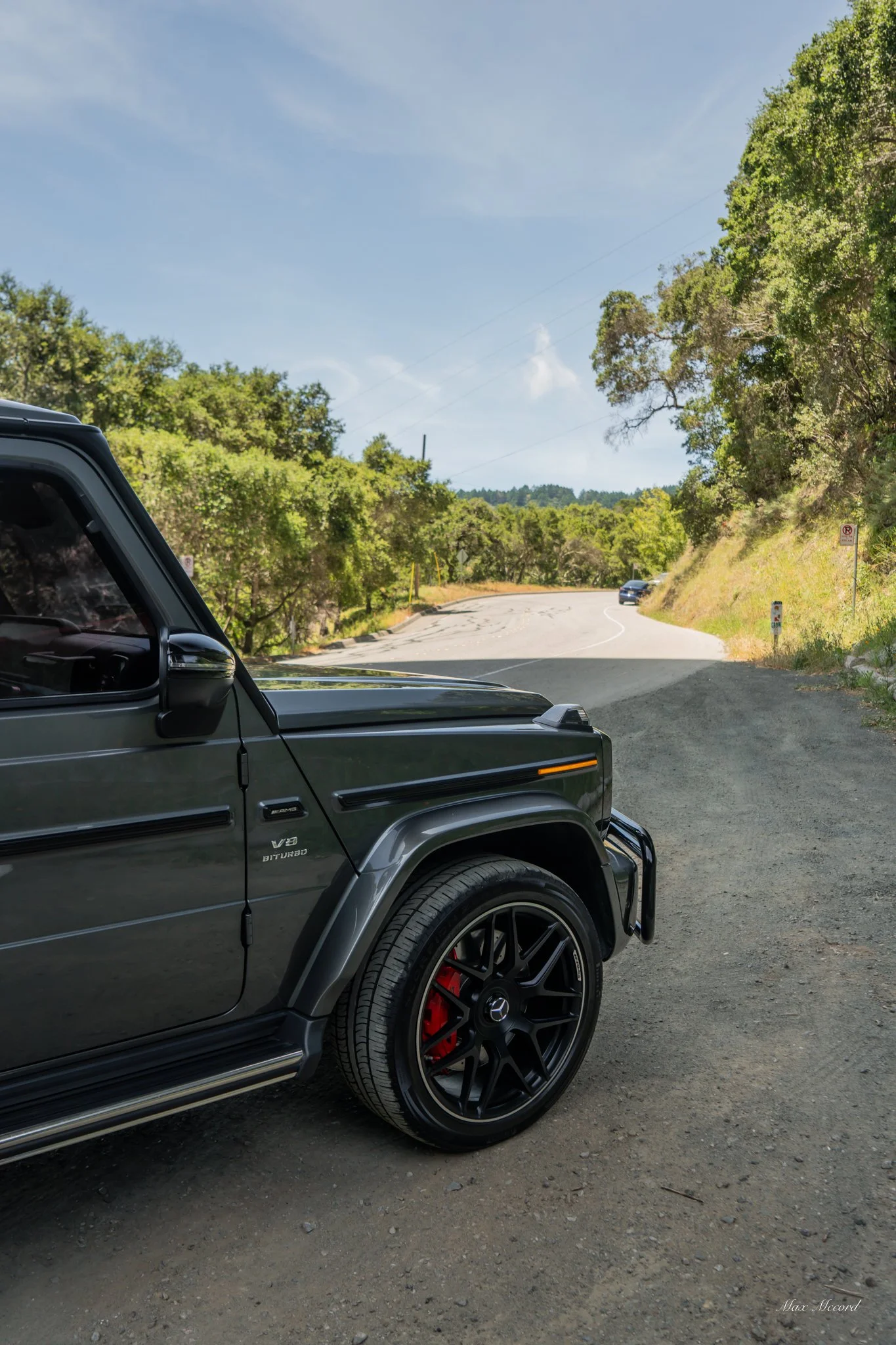 Close-up of the front part of a black Mercedes-Benz G-Class SUV parked on a dirt roadside, with a winding paved road and green trees in the background under a partly cloudy sky.