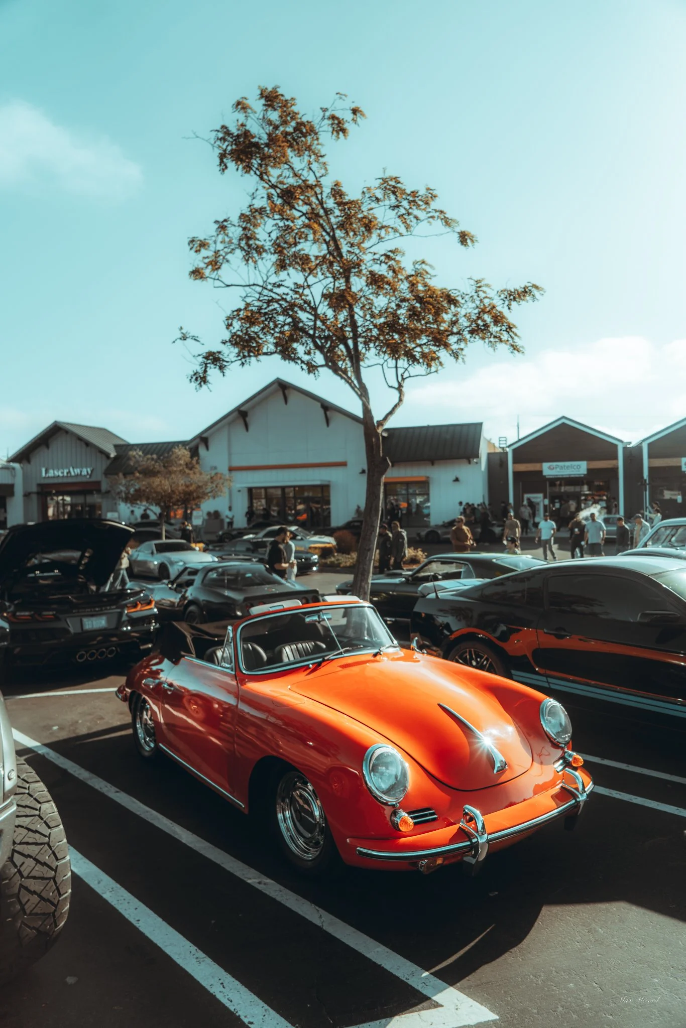 A bright orange vintage sports car parked in a lot with several modern cars around it, with a shopping center in the background and a tree in the foreground.
