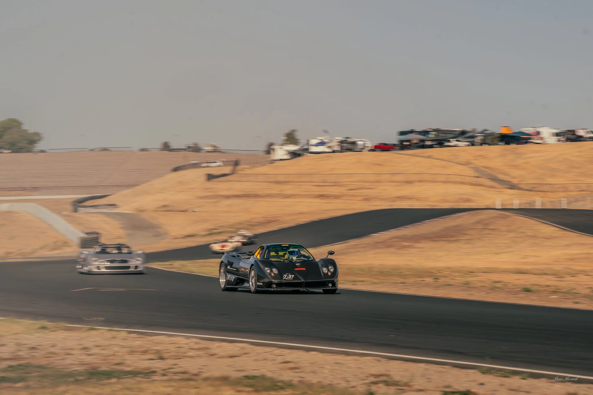 A black race car with yellow accents on a race track with a clear sky, followed by a silver race car, and a line of parked cars and trailers on a hill in the background.