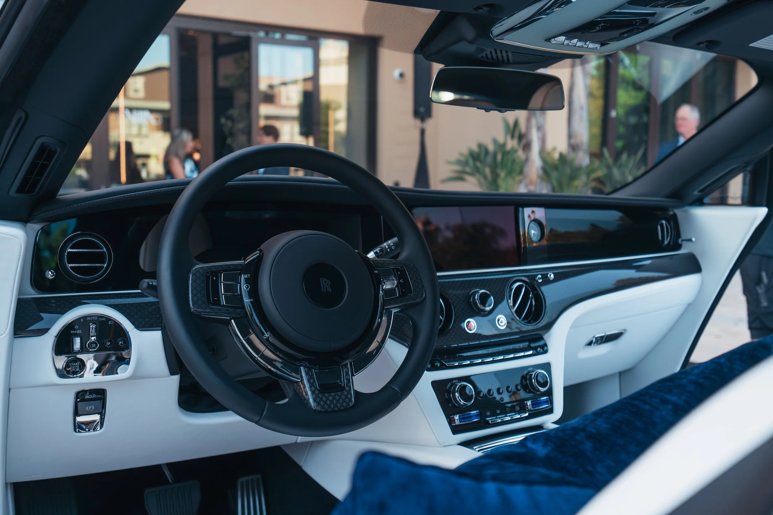 Interior of a luxury car showing a black steering wheel with the Rolls-Royce logo, dashboard with multiple controls, and a large digital display, outside view visible through the windshield.