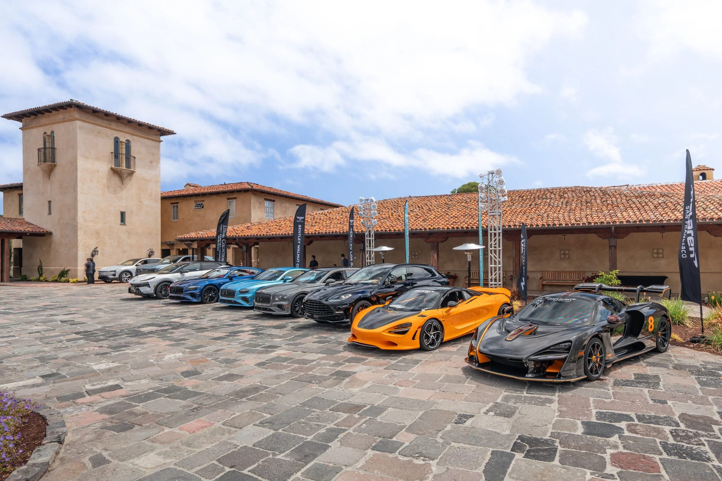 A lineup of ten luxury sports cars parked in a courtyard with stone paving, in front of a building with a tiled roof and beige walls, under a partly cloudy sky. Flags with car brand logos are visible.
