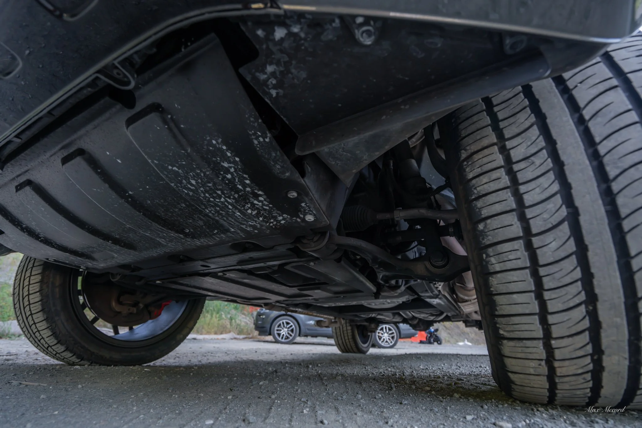 Underneath view of a vehicle showing the oil pan, suspension components, and tire tread on a gravel surface with other cars in the background.