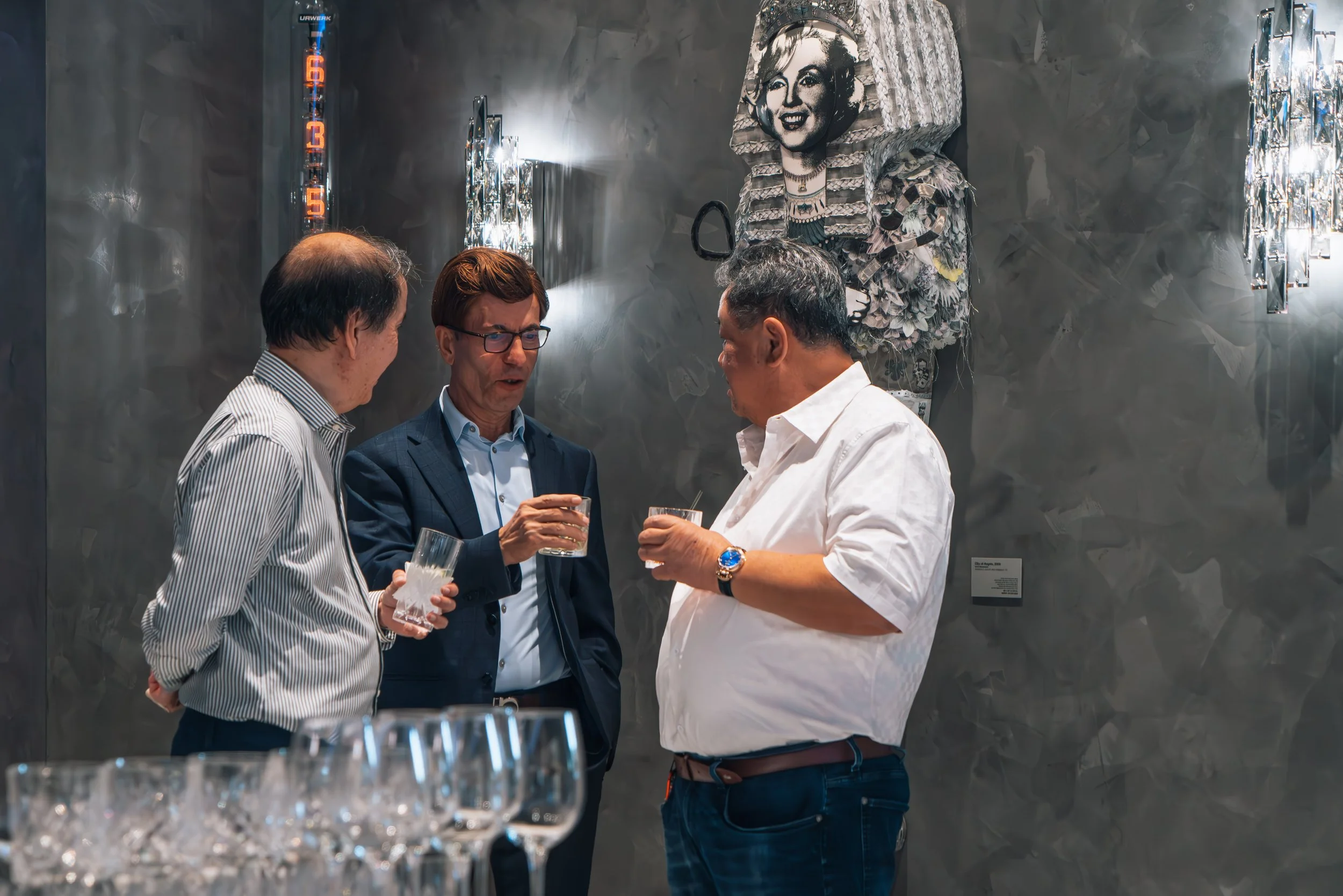 Three men in business attire having drinks and engaging in conversation at an art gallery or exhibition with modern artwork on the wall behind them
