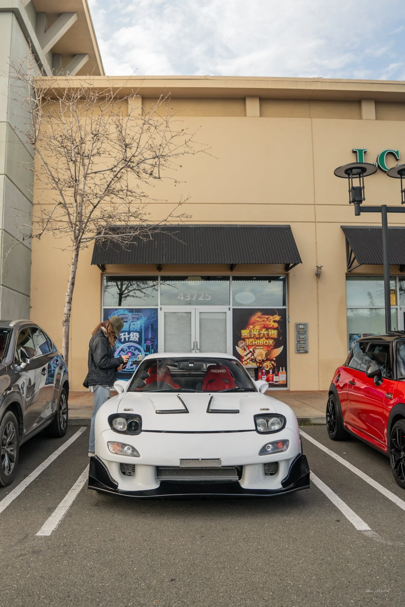 A white sports car with black accents and red interior parked in a parking lot in front of a beige commercial building, with a person standing beside it holding a phone.