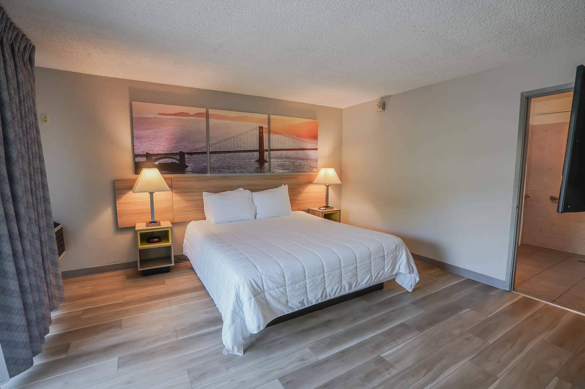A hotel room with a bed, two bedside lamps, artwork of the San Francisco bridge at sunset on the wall, beige walls, wooden flooring, and an open doorway to a bathroom.