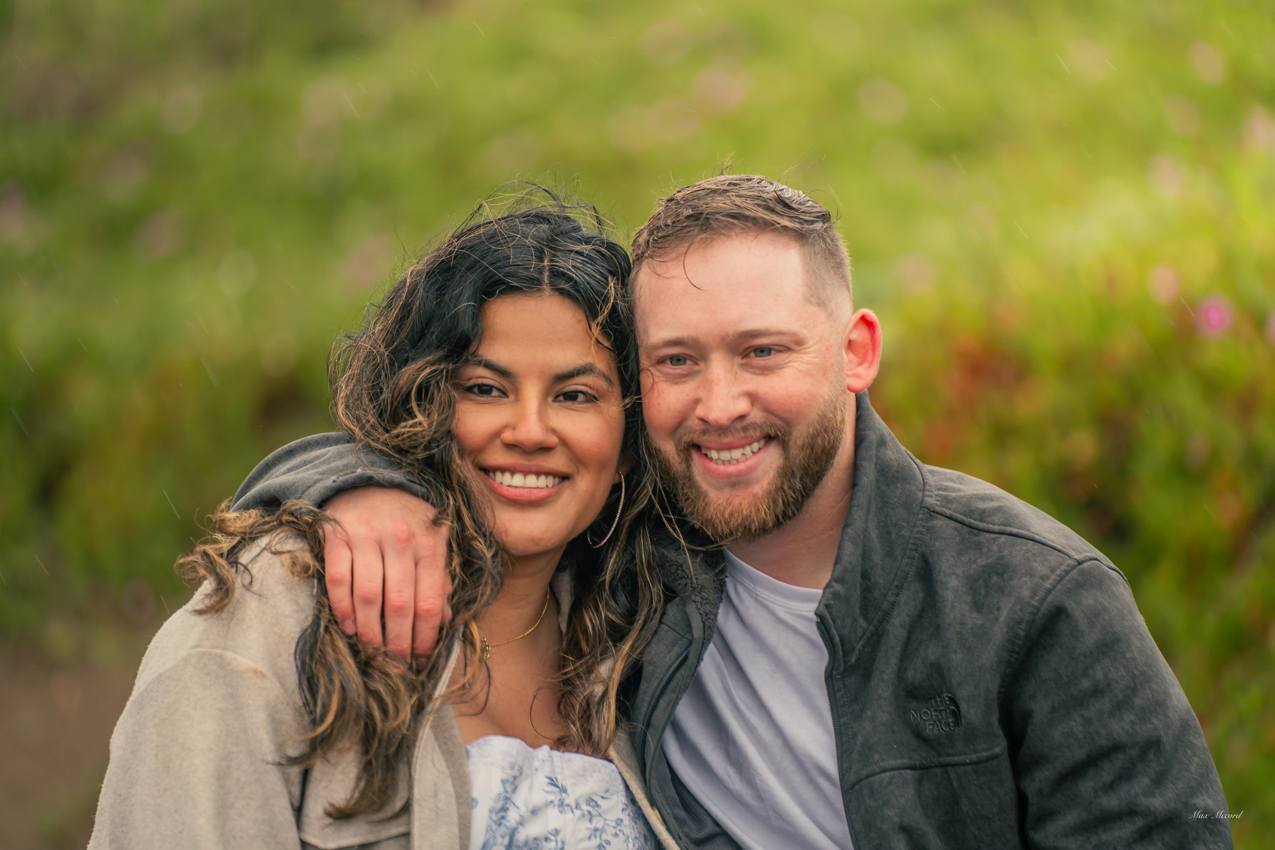 A smiling couple, a woman with long, curly dark hair and a man with a beard, are standing close together outdoors with a green, blurred background, and the woman has her arm around the man's shoulder.