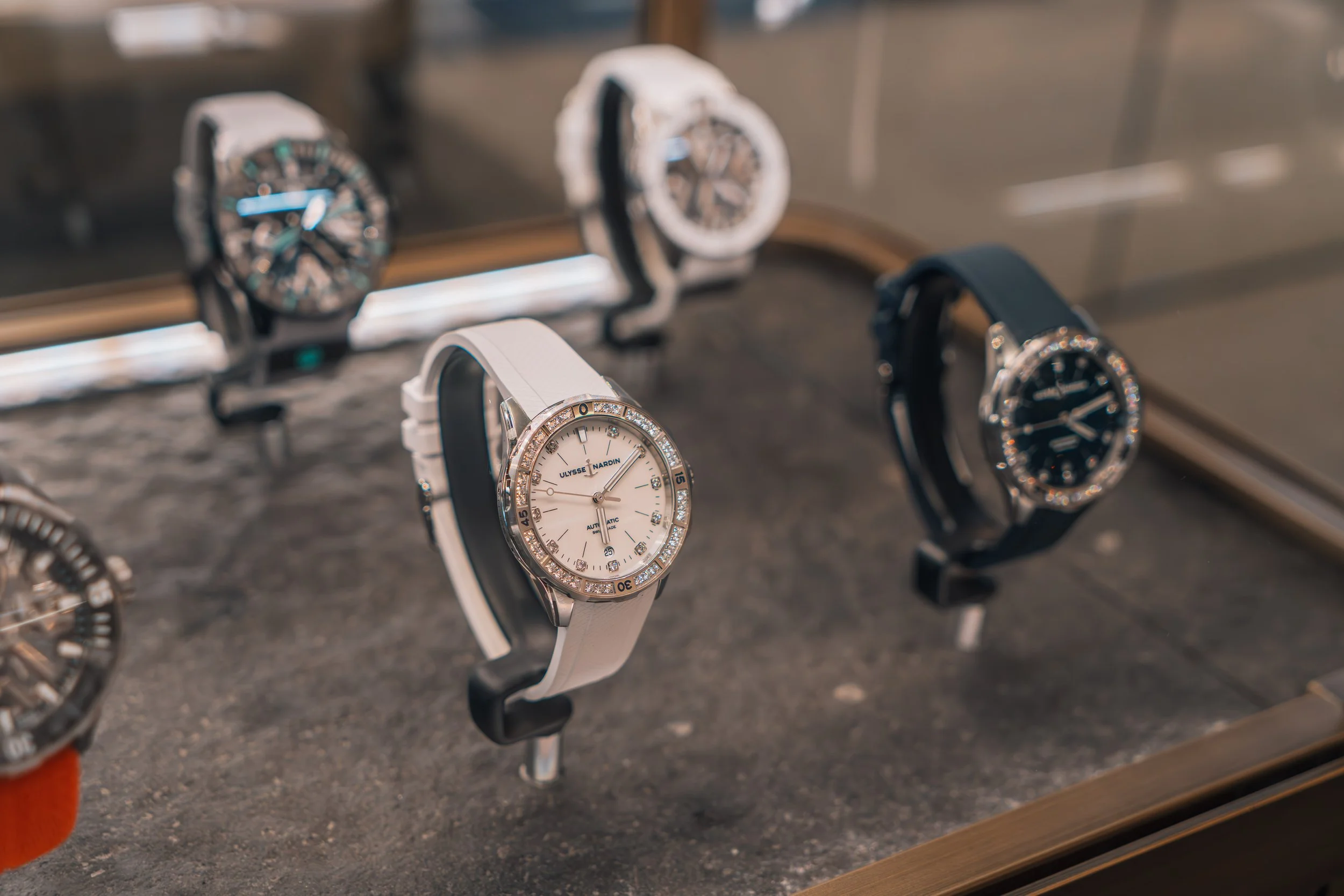 Display case with luxury wristwatches, including a white strap watch with diamond accents, a black strap watch, and a white strap watch in the background.