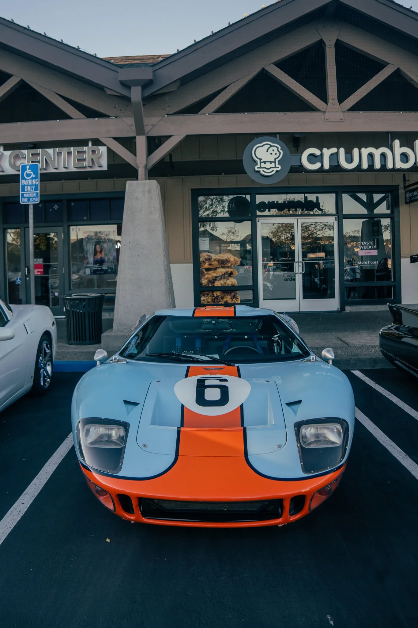 A classic Ford GT race car with a light blue and orange stripe design, featuring the number 6, parked in front of a Crumbl cookie store.