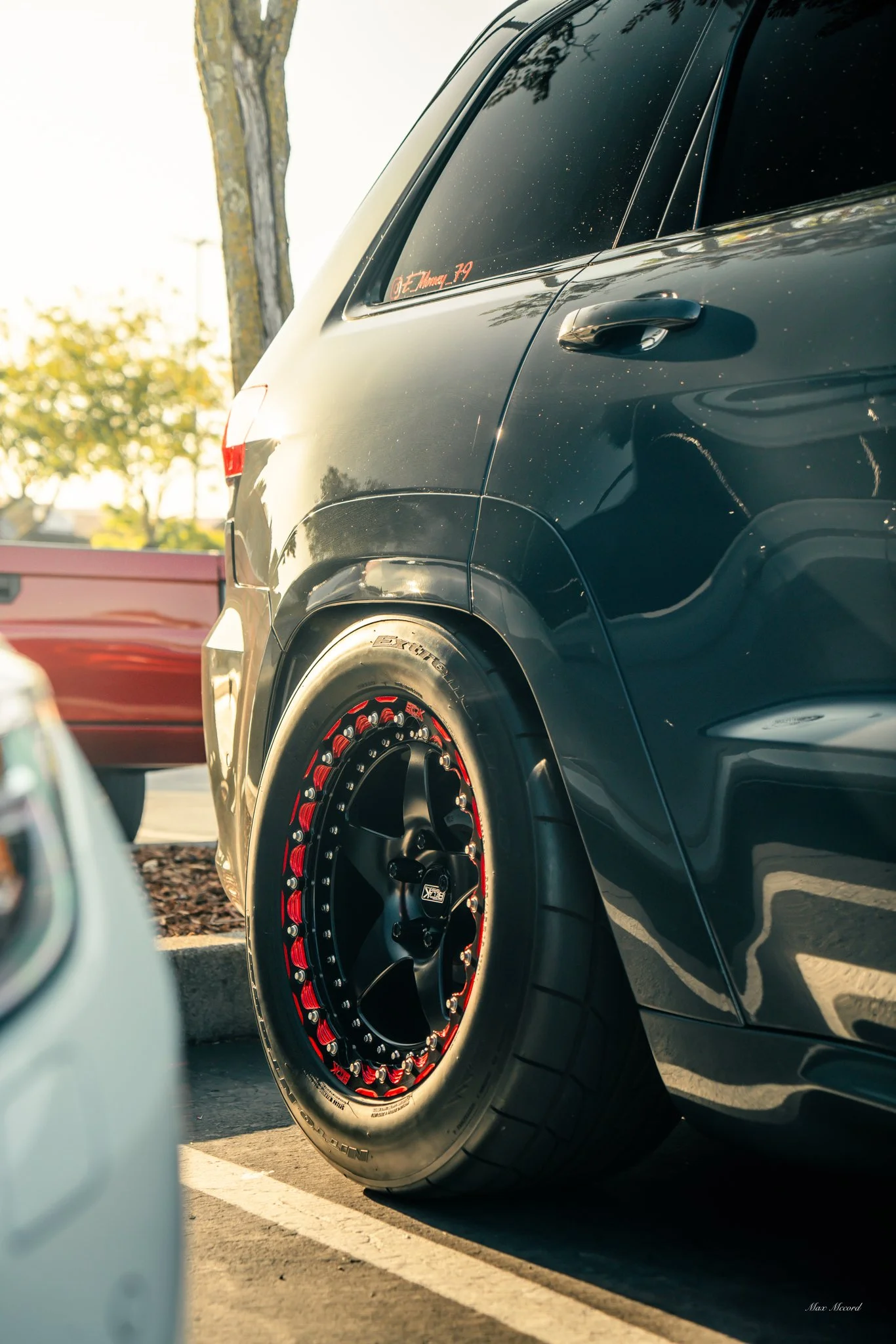 Close-up of a dark-colored car's rear wheel with custom red and black rims parked in a lot, with trees and another vehicle in the background.