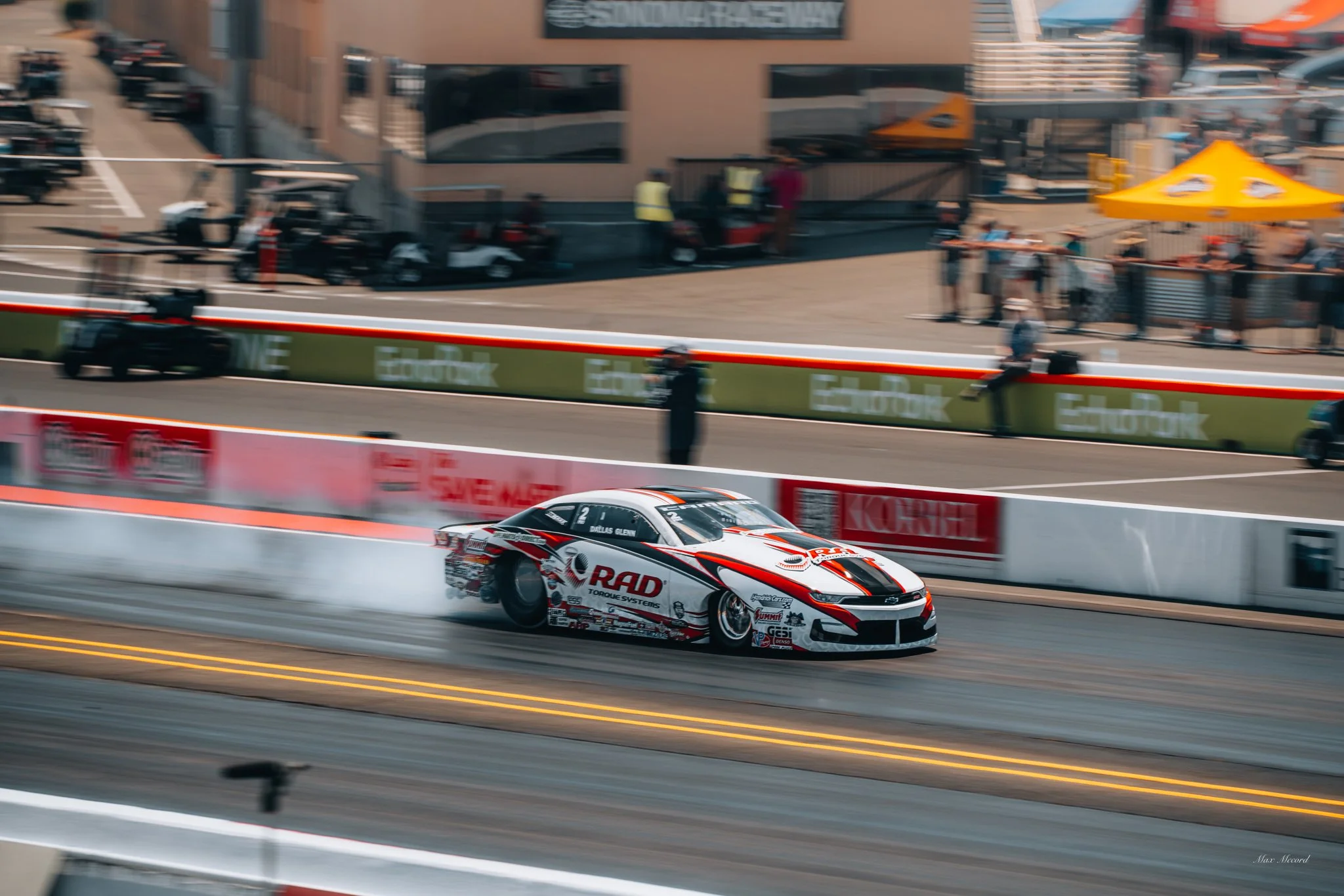 A race car during a drag race, emitting smoke from its tires, on a racing track with spectators and officials in the background.