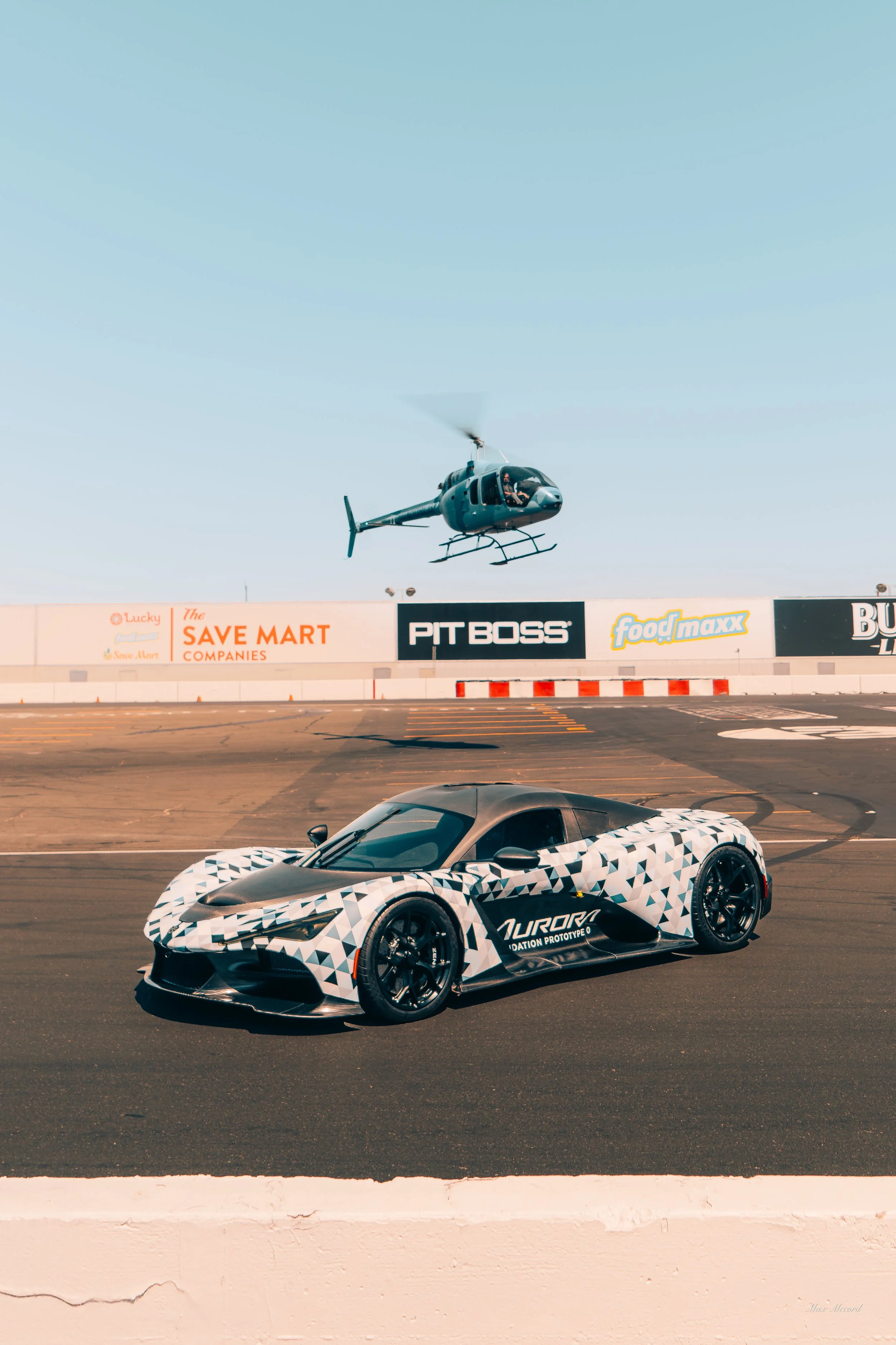 A camouflaged prototype sports car parked on a racetrack with a helicopter flying overhead.
