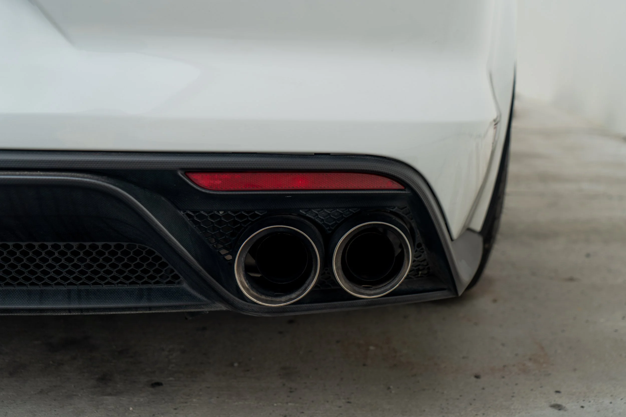 Close-up of the rear end of a white sports car, showing dual exhaust pipes, black rear diffuser, and a red reflector.
