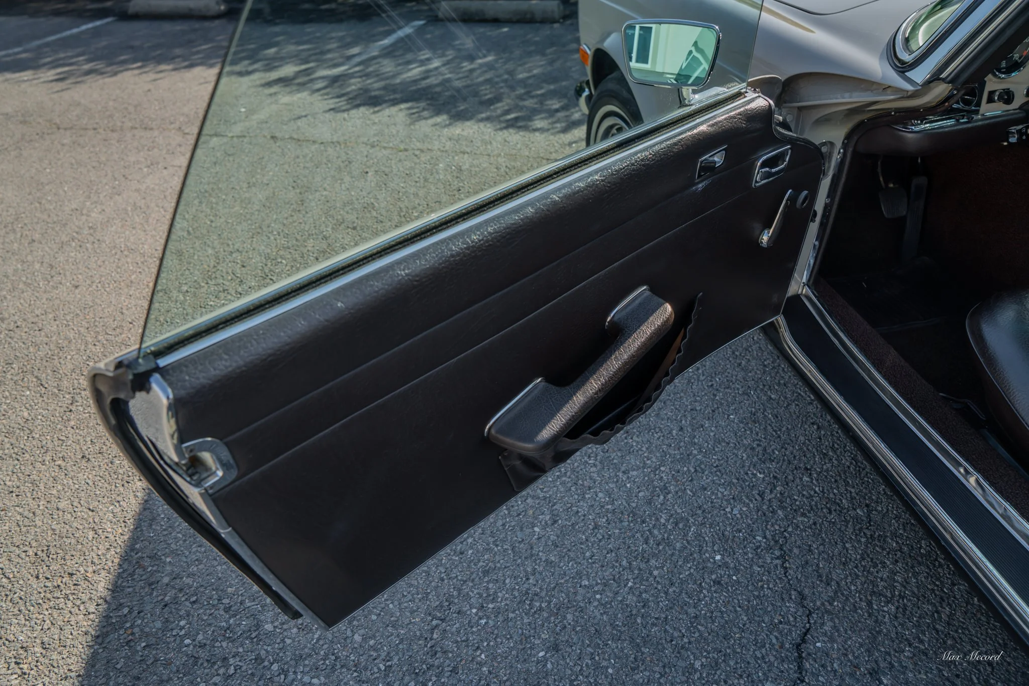 Open car door showing interior panel, handle, and window, with a view of the outside sidewalk and a parked silver car.