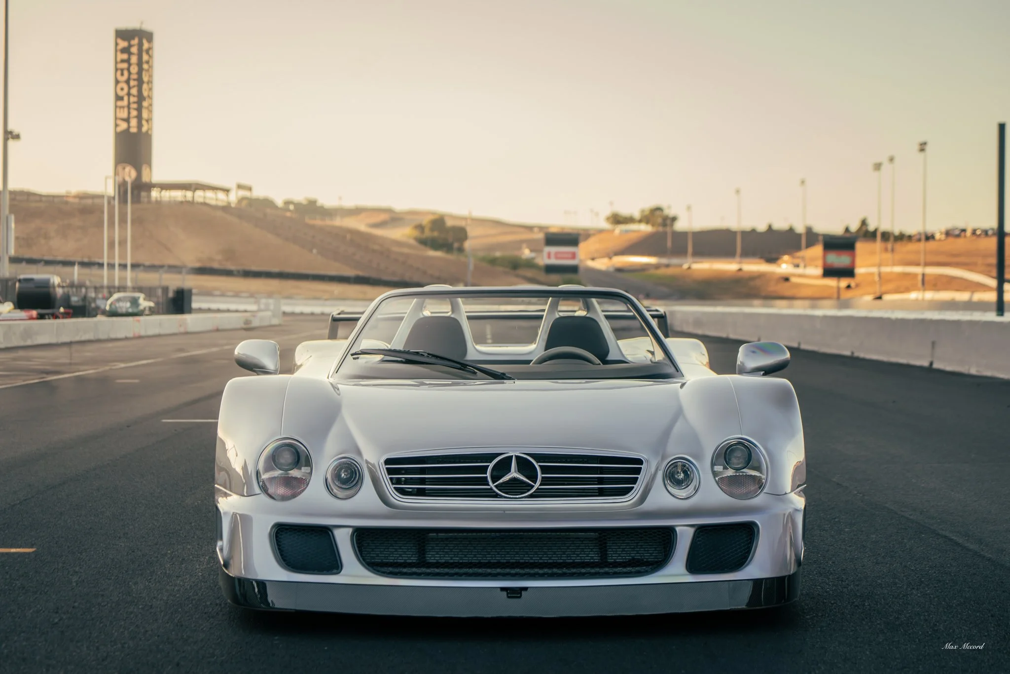 A white Mercedes-Benz race car on a race track with hills and signs in the background during daytime.