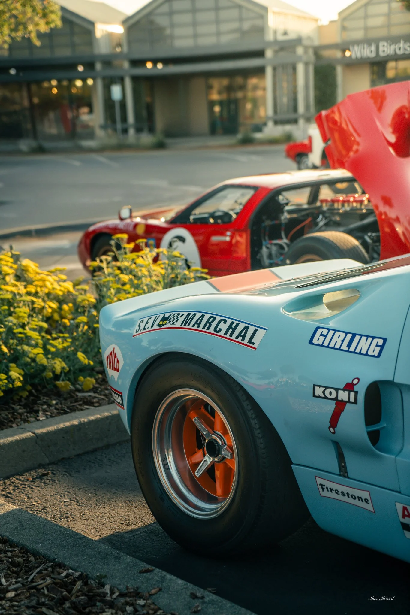Close-up of vintage race cars parked outside, with the front wheels and body panels visible, featuring logos and sponsor decals like S.E. Marchal, GIRLING, KONI, Firestone, and others.
