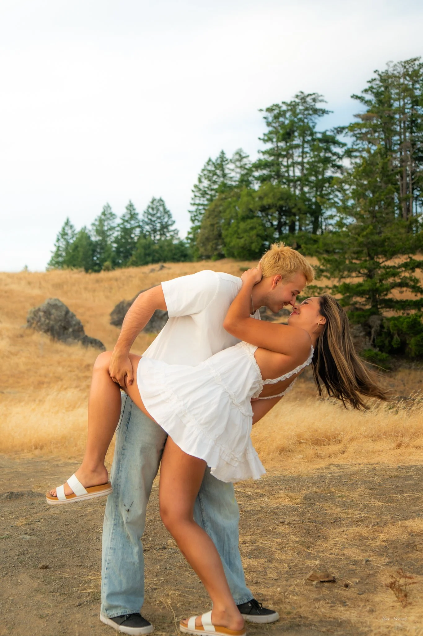 A couple dancing outdoors on a dirt field with trees in the background, the man is holding the woman and they are about to kiss.